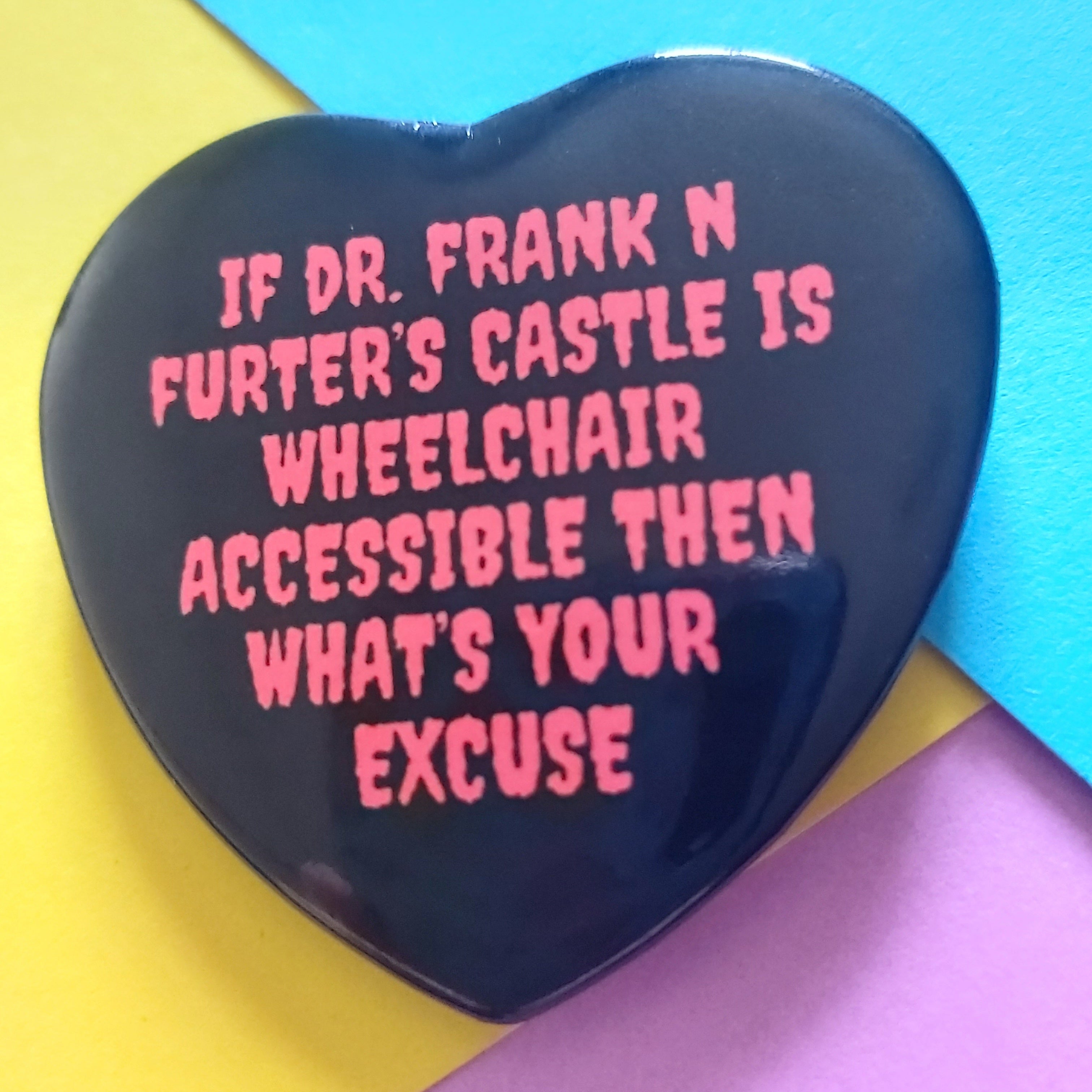 A heart-shaped black pin with red text reading 'IF DR. FRANK N FURTER'S CASTLE IS WHEELCHAIR ACCESSIBLE THEN WHAT'S YOUR EXCUSE', displayed on a colourful background of yellow, blue, and pink paper.