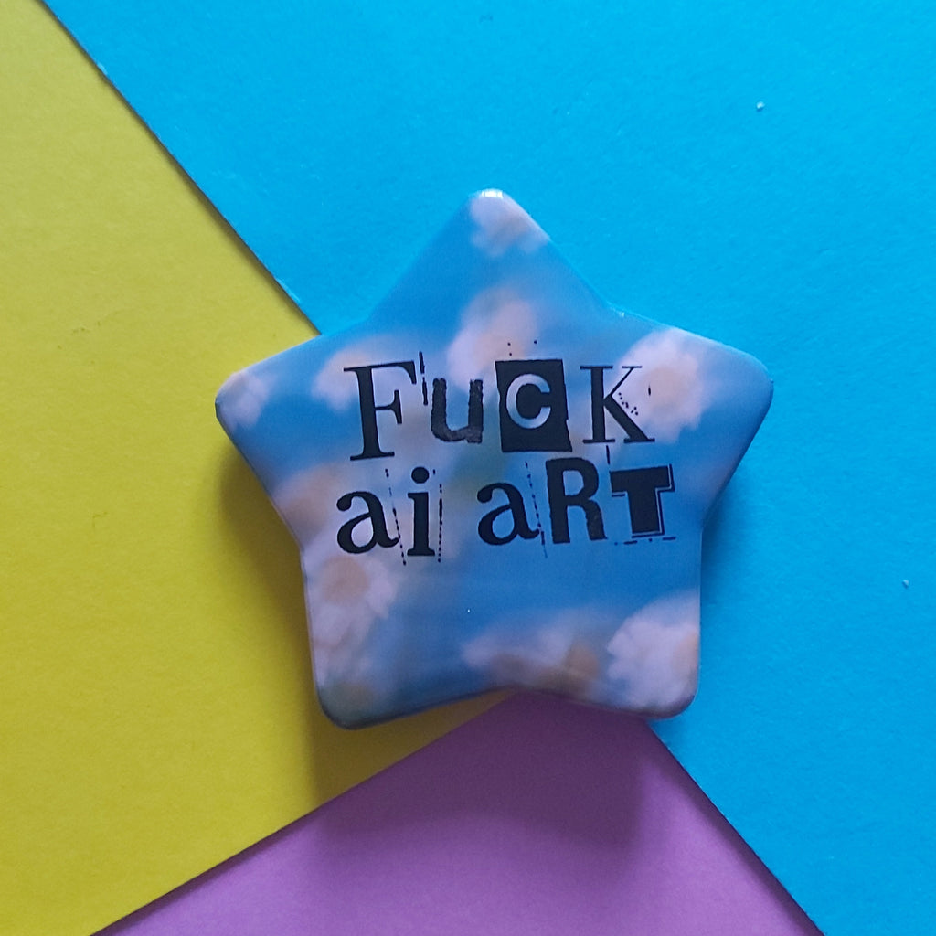 A star-shaped pinback button with a blue sky and white clouds pattern, featuring the text 'Fuck AI art' in black. The pin is displayed on a colourful background composed of blue, yellow, and pink paper sheets.