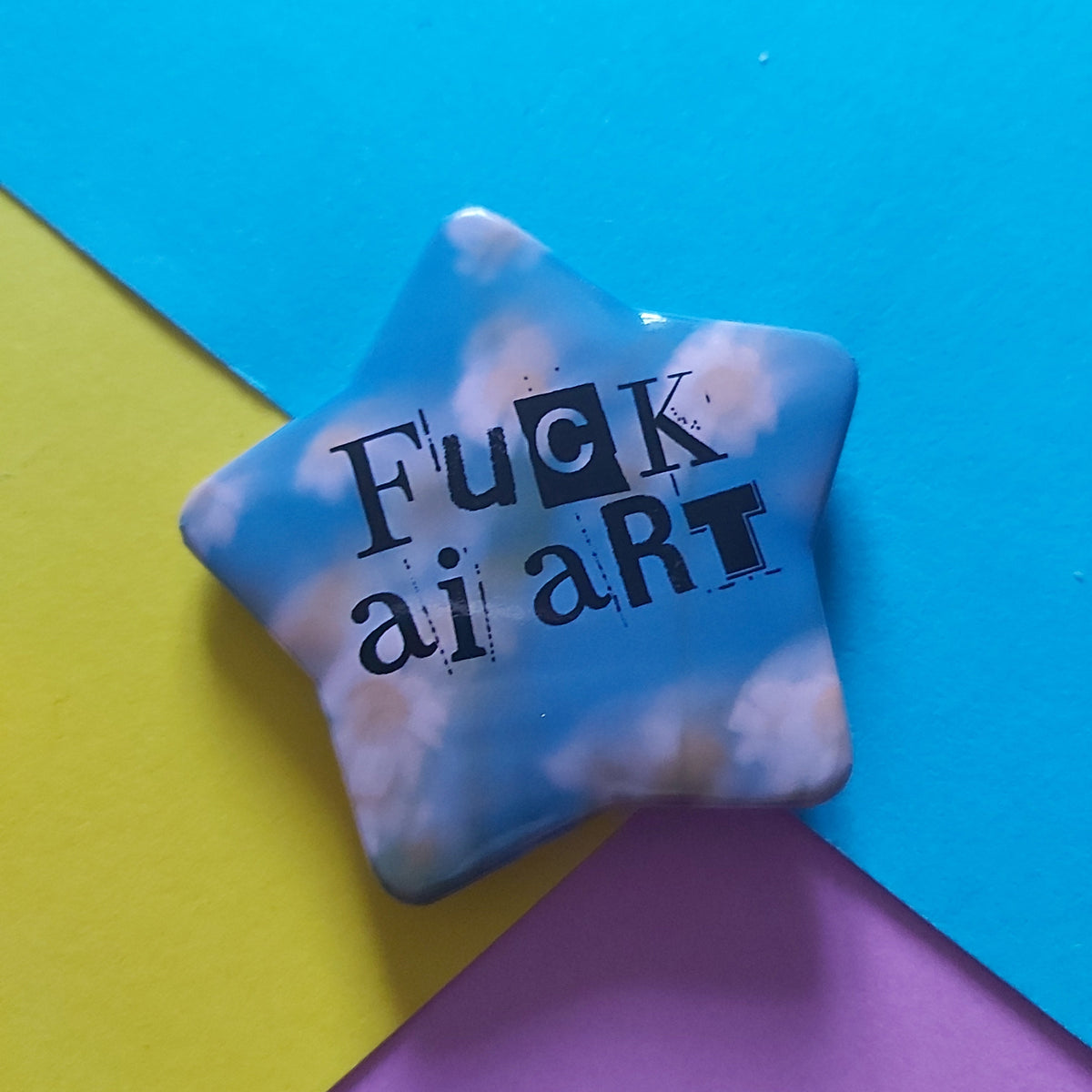 A star-shaped acrylic pin with a blue sky and white clouds pattern, featuring the text 'Fuck AI art' in black. The pin is displayed on a background of three colored paper sheets: blue, yellow, and pink.