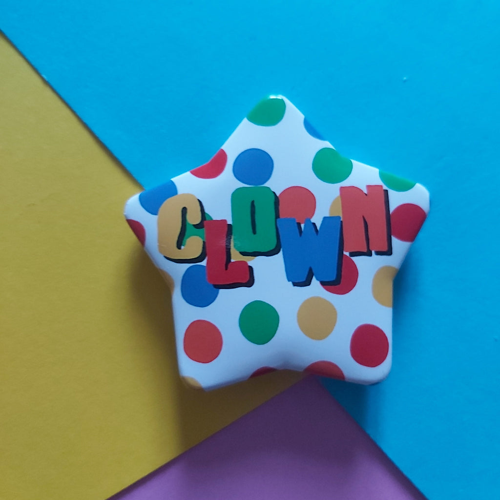 A star-shaped badge with a white background, colourful polka dots, and the word 'CLOWN' spelled out in raised multicoloured letters, photographed on a geometric multicolour background of blue, yellow, and purple paper.