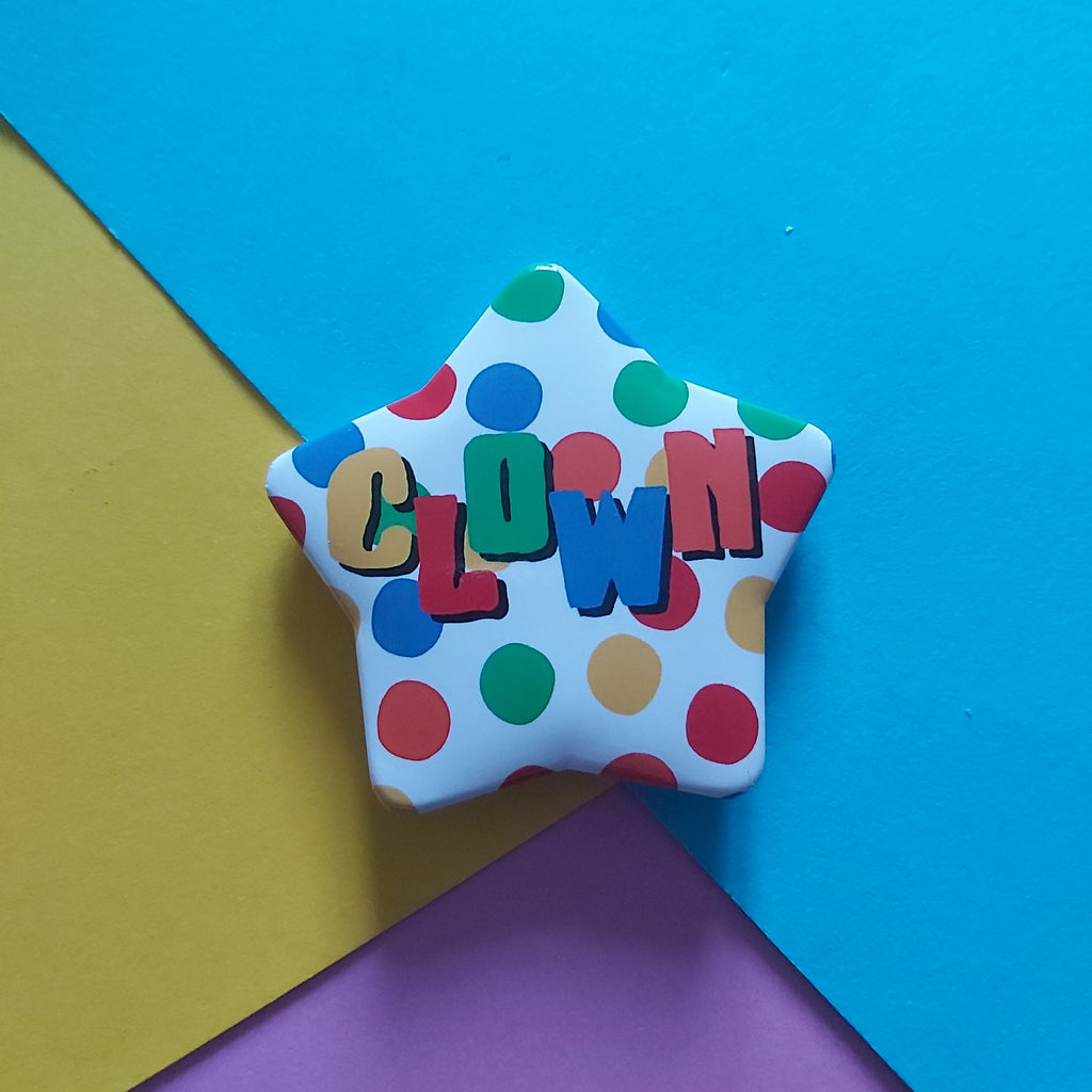 A star-shaped badge with a white background, multicoloured polka dots, and the word 'CLOWN' spelled out in colourful letters, set against a geometric background of blue, yellow, and purple paper.