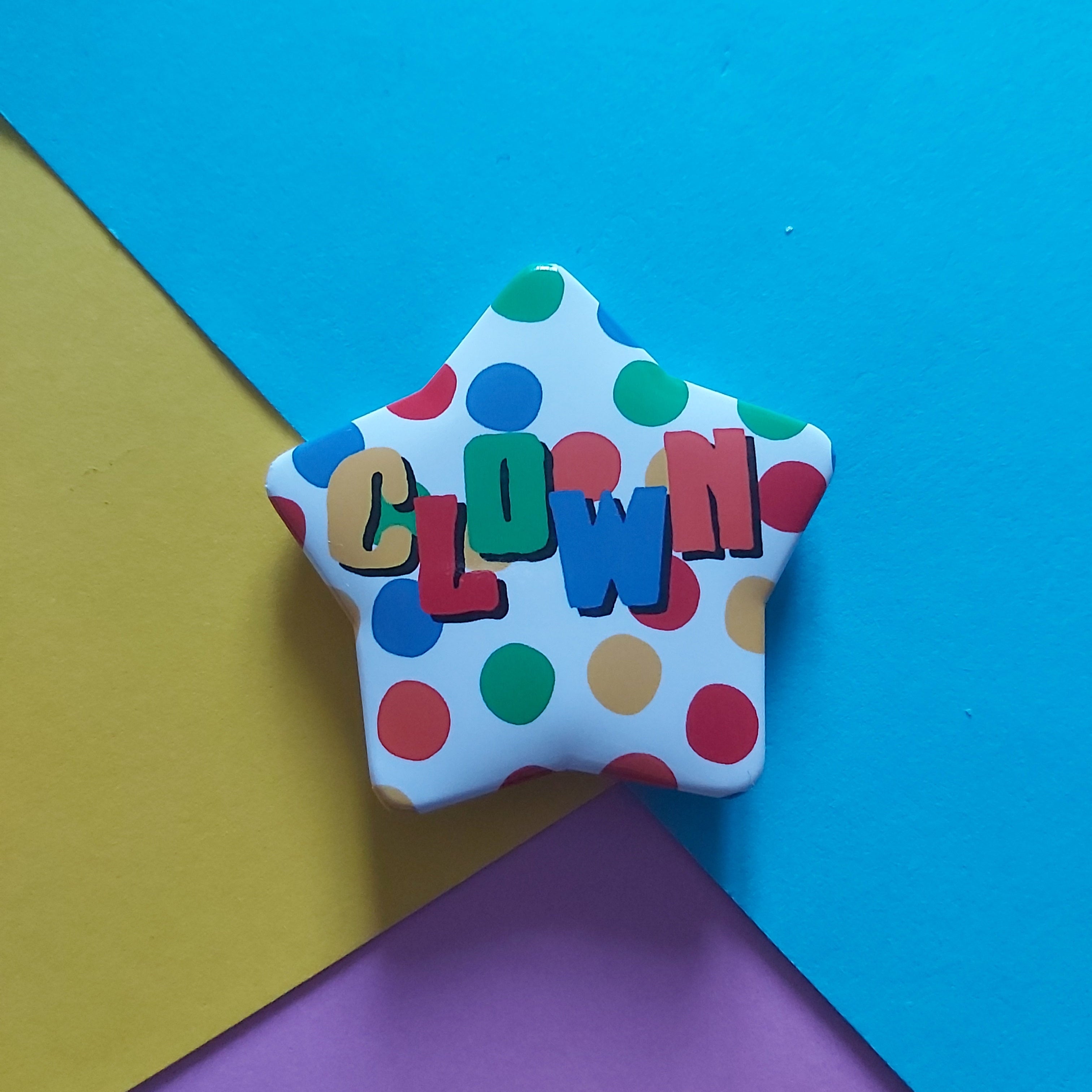 A star-shaped badge with a white background, multicoloured polka dots, and the word 'CLOWN' spelled out in colourful letters, set against a geometric background of blue, yellow, and purple paper.