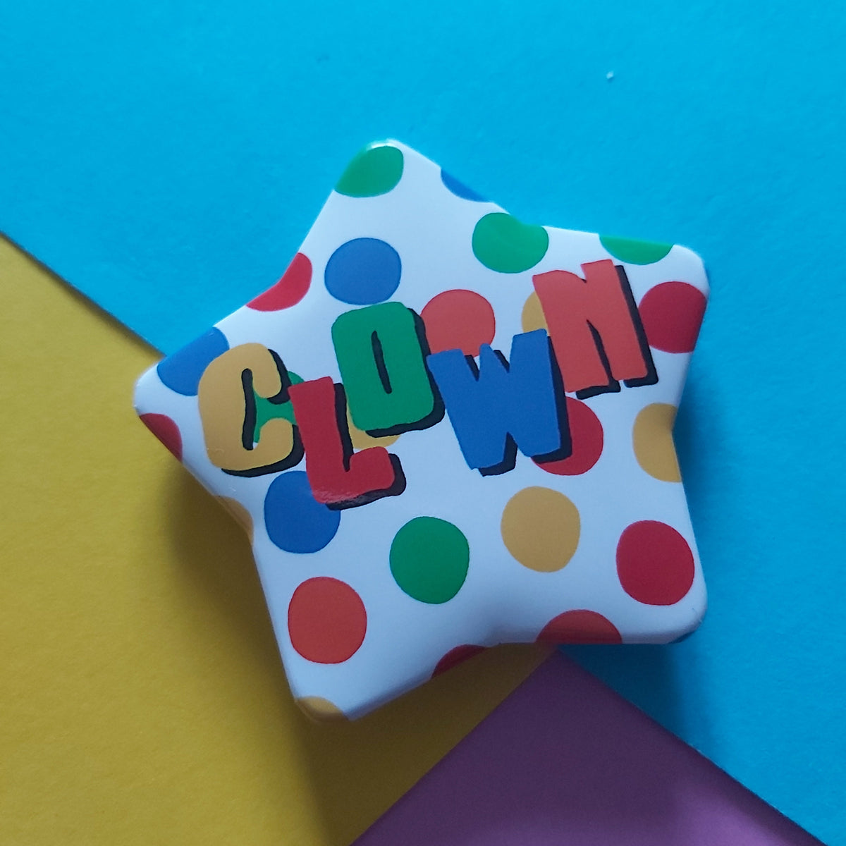 A star-shaped badge with colourful clown polka dots and raised multicoloured letters spelling 'CLOWN', photographed on a bright blue background with yellow and purple paper sections.