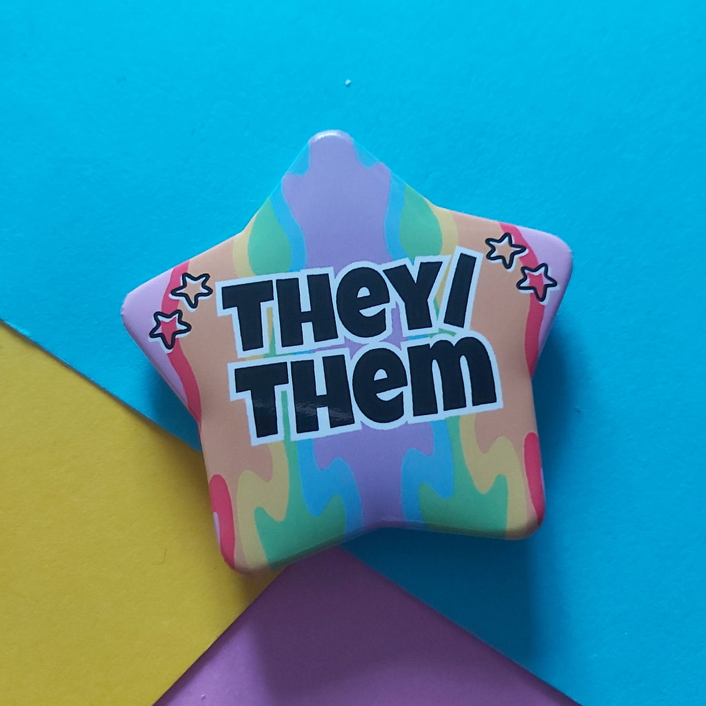 A star-shaped pin with the text 'THEY/ THEM' in bold black letters, set against a pastel rainbow tie-dye pattern. The pin is photographed on a colourful background composed of blue, yellow, and purple paper sheets.