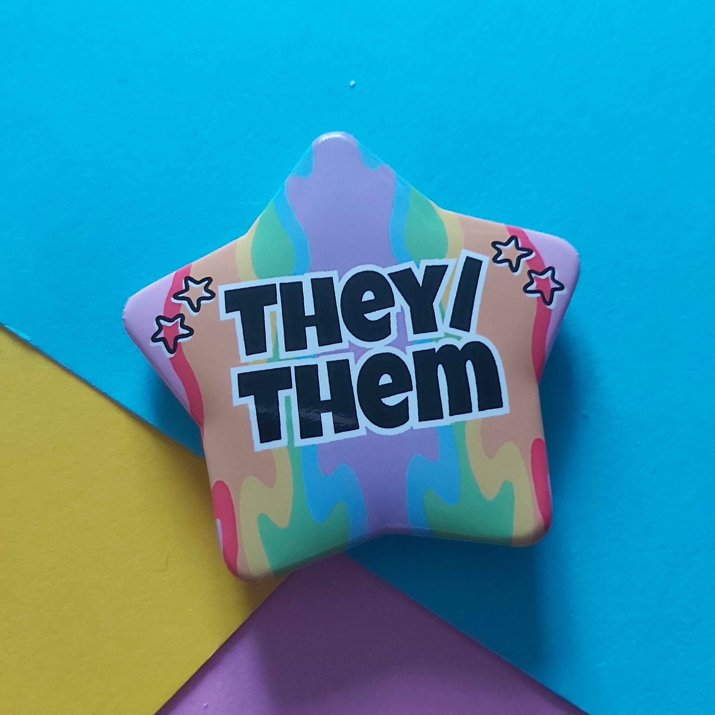 A star-shaped pin with the text 'THEY/ THEM' in bold black letters, set against a pastel rainbow tie-dye pattern. The pin is photographed on a colourful background composed of blue, yellow, and purple paper sheets.