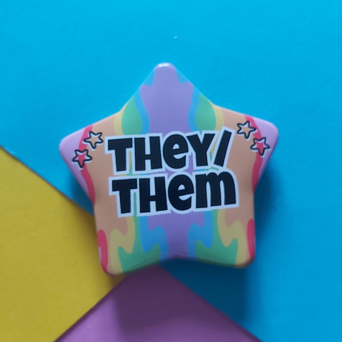 A star-shaped pin with a colourful tie-dye pattern and the text 'THEY/ THEM' in bold black letters, set against a background of bright blue, yellow, and purple paper.