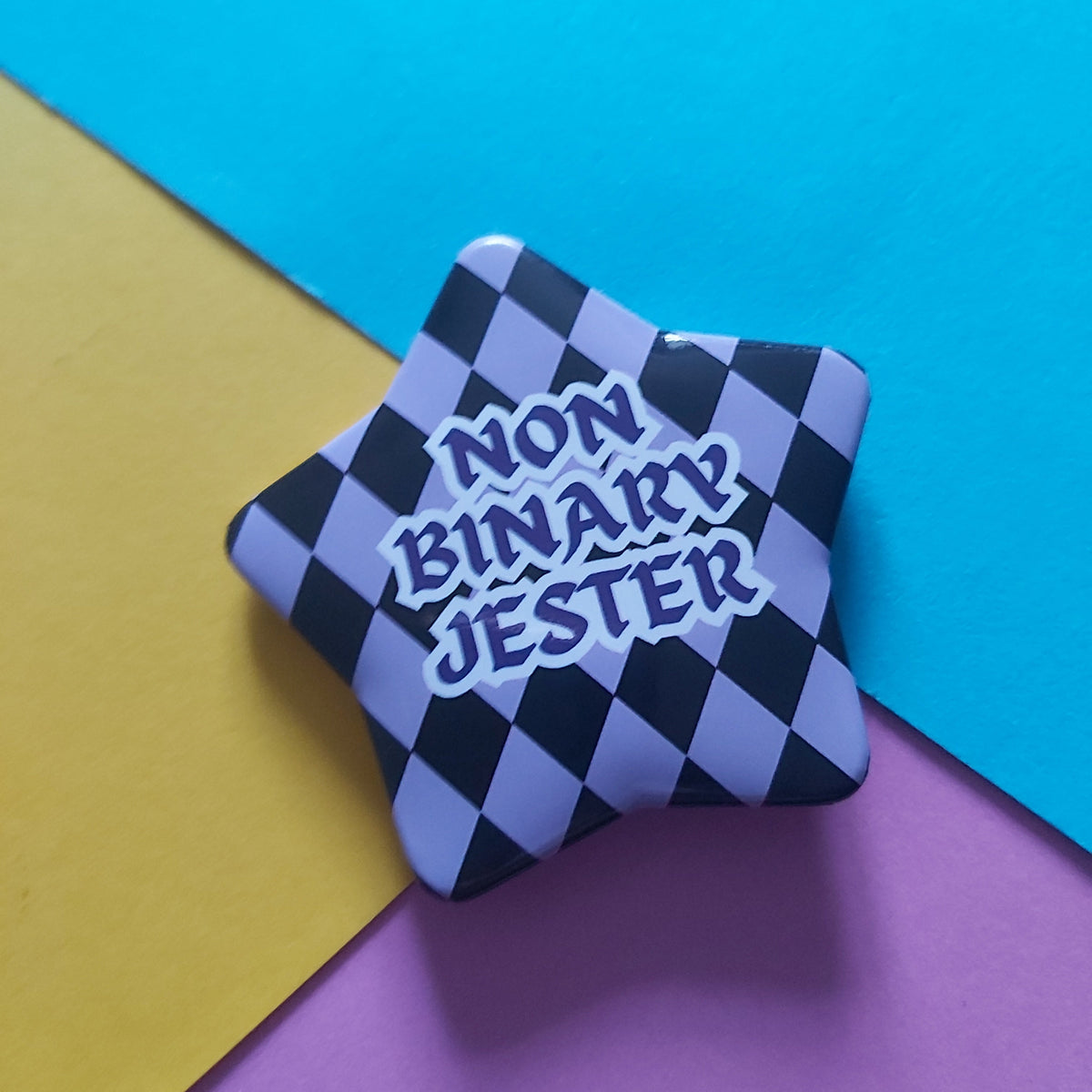 A star-shaped pin badge with a black and lavender-purple checkered pattern and the text 'NON BINARY JESTER' in stylized lettering, displayed on a diagonally split yellow, blue, and purple background.