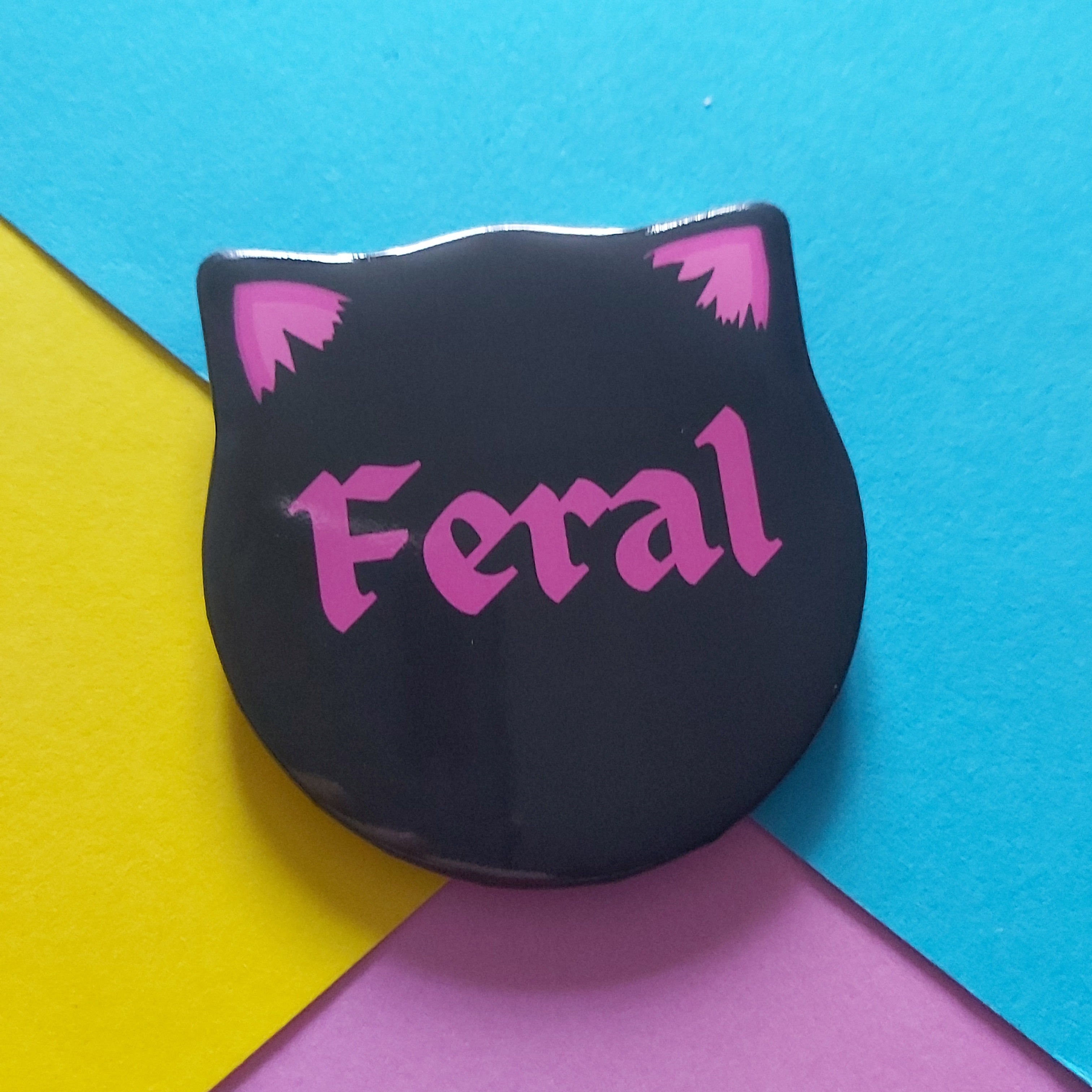 A black cat-head-shaped pin badge with pink cat ears and the word 'Feral' in pink gothic lettering, placed on a colourful background split into blue, yellow, and purple sections.