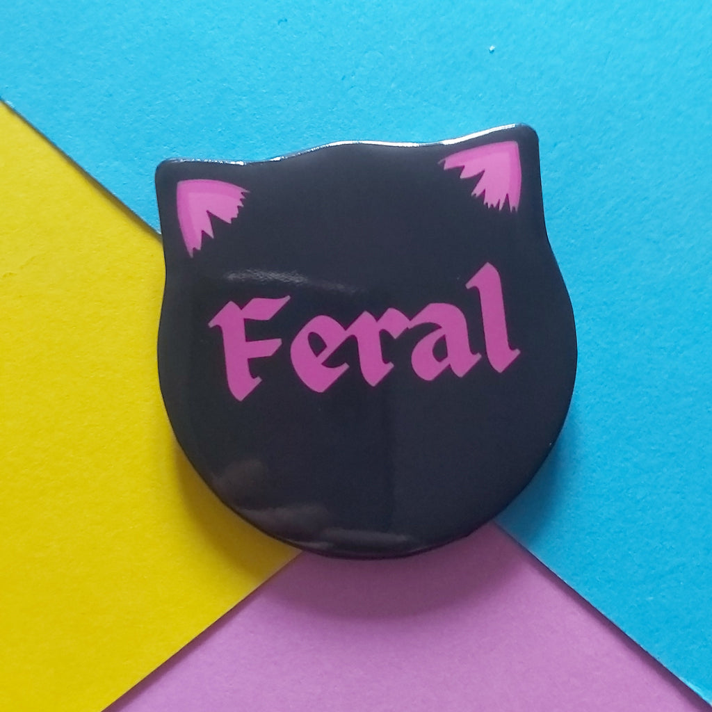 A black cat-head-shaped pin badge with pink ears and the word 'Feral' in pink gothic lettering, placed on a colourful background of blue, yellow, and purple paper.
