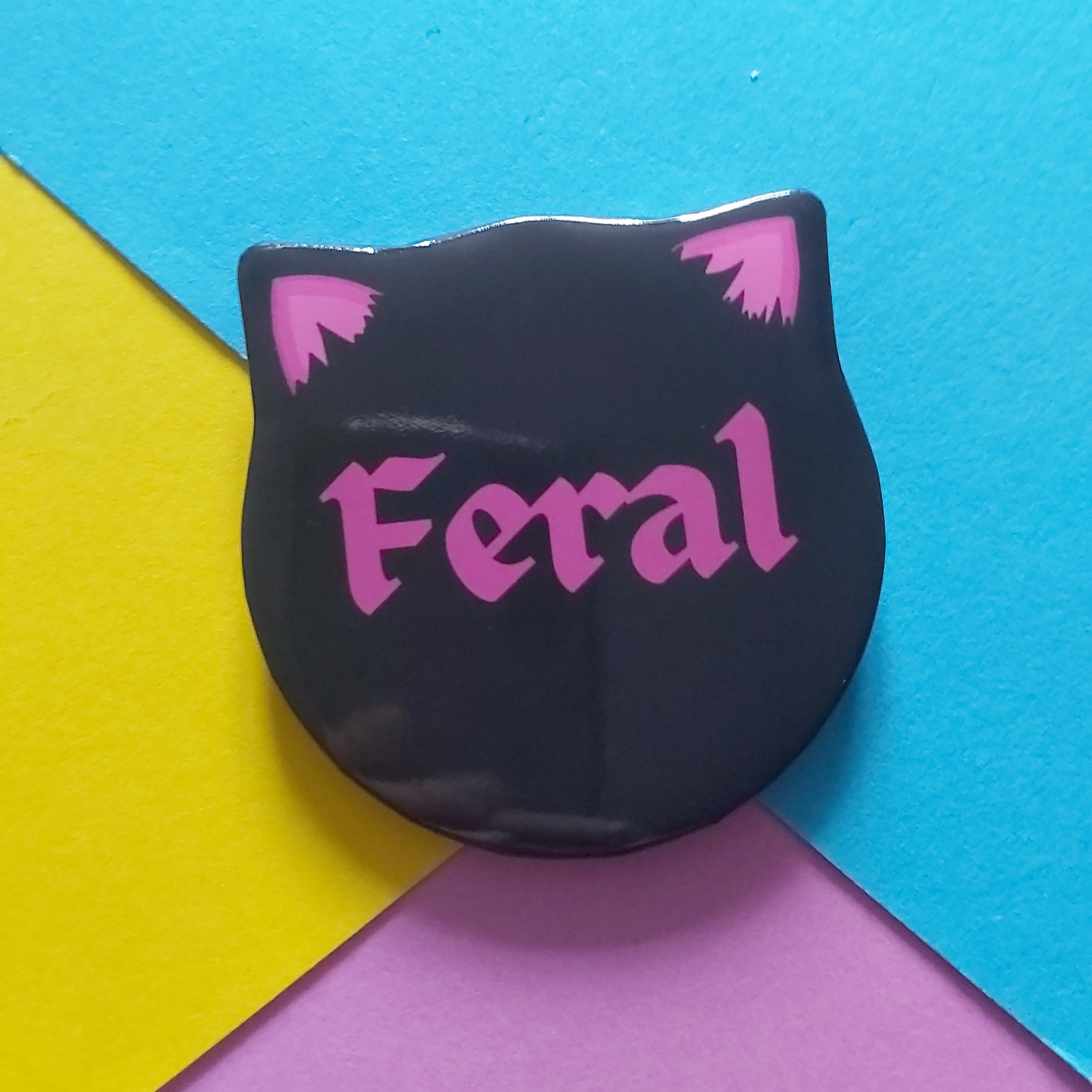 A black cat-head-shaped pin badge with pink ears and the word 'Feral' in pink gothic lettering, placed on a colourful background of blue, yellow, and purple paper.