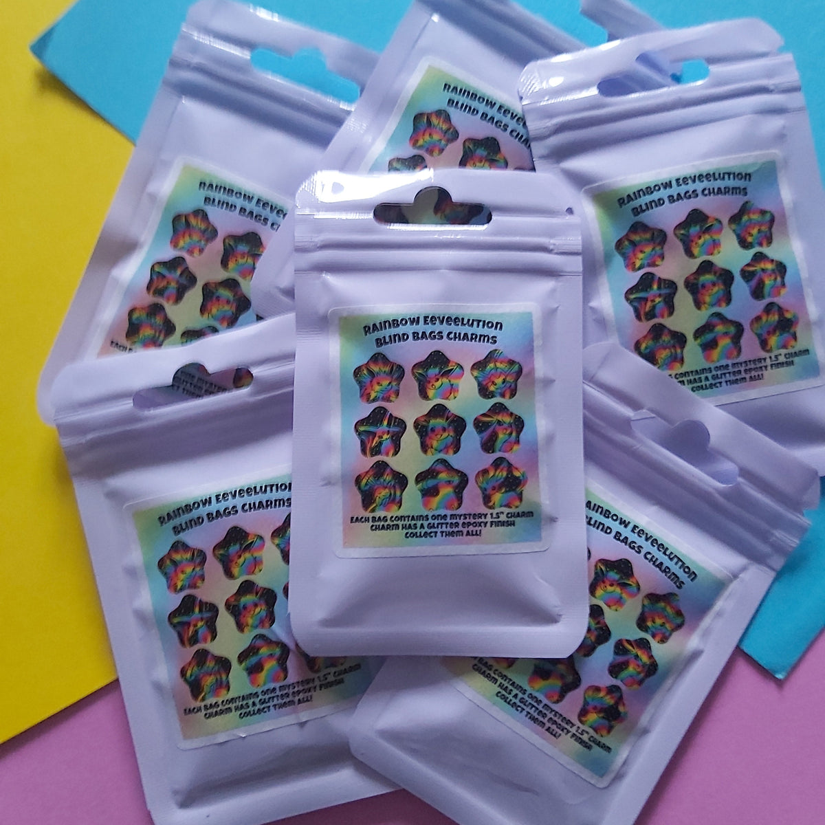 Five small white resealable pouches with colourful labels are arranged on a bright pink and blue background. The labels show the text 'Rainbow Eeveelution Blind Bag Charms' and depict multicoloured starburst charms.