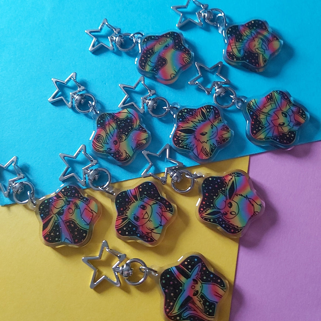 Nine silver keychains with star-shaped links and holographic rainbow pendants are arranged on a split yellow and blue background with a purple corner.
