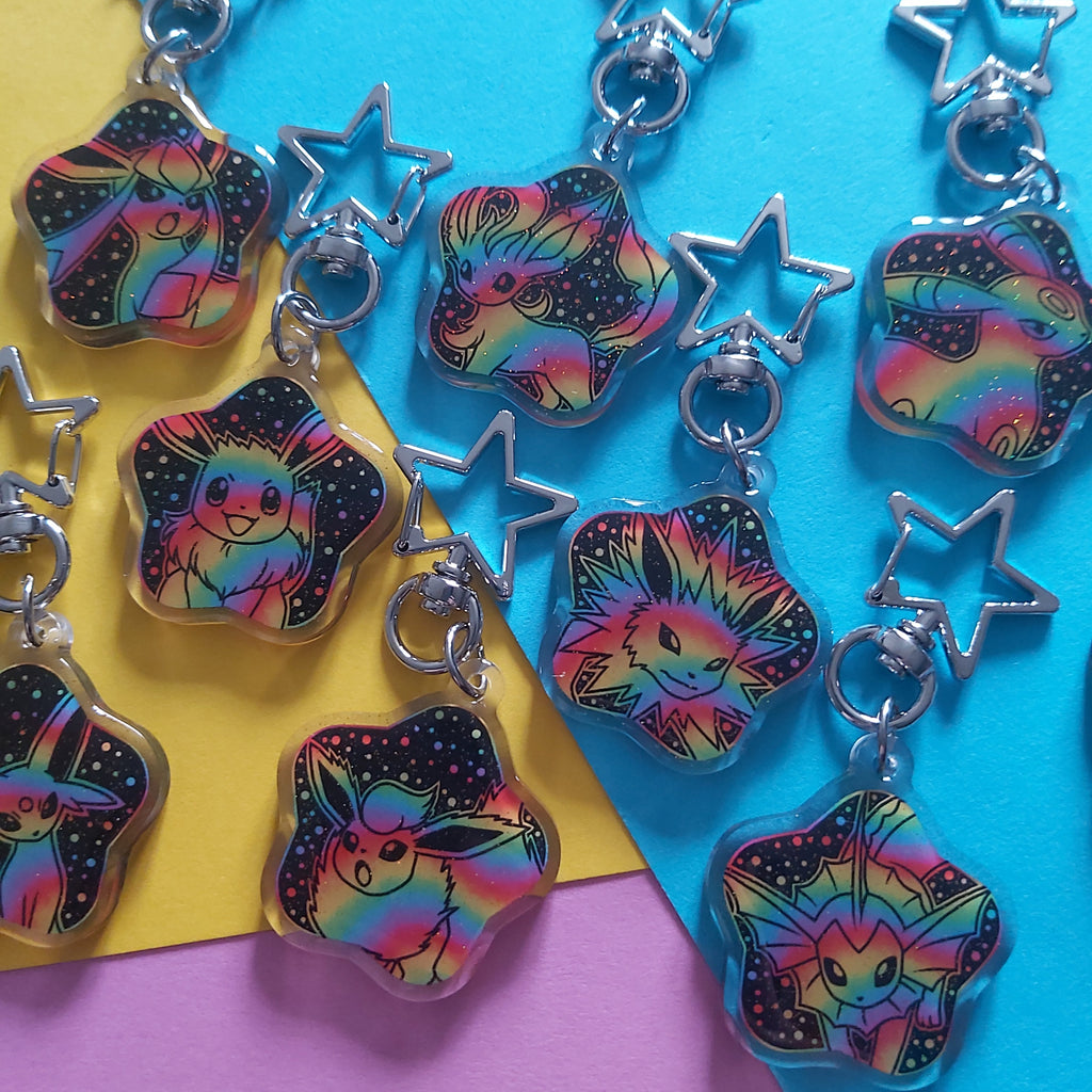 A set of star-shaped keychains featuring colorful, iridescent rainbow designs of cartoon creatures on a black starry background, attached to silver metal rings with star charms, arranged on a diagonally split yellow, blue, and purple paper background.