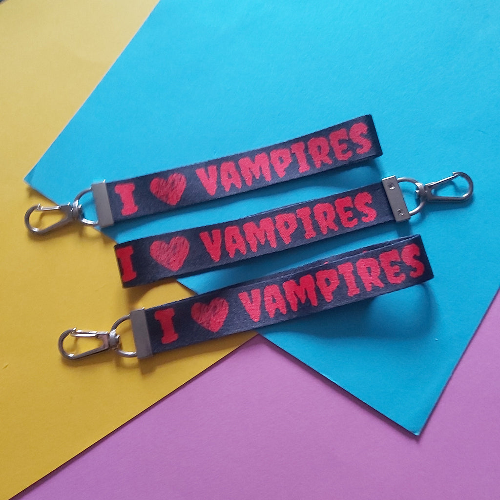 Three black fabric keychains with silver metal clamps, printed with 'I ♥ VAMPIRES' in red, arranged on a background of yellow, blue, and pink paper.