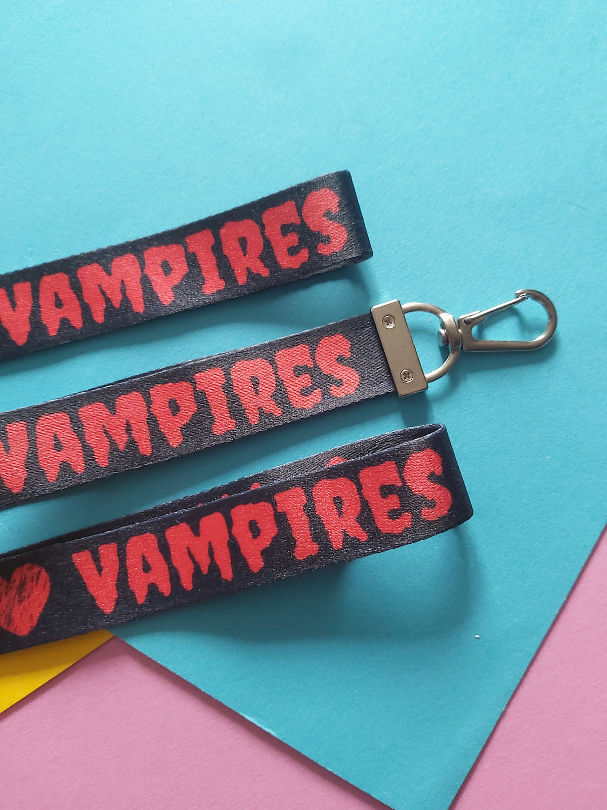 Three black fabric wristlets with red 'VAMPIRES' text and silver clasp hardware, arranged diagonally on a colorful background of blue, pink, and yellow paper.