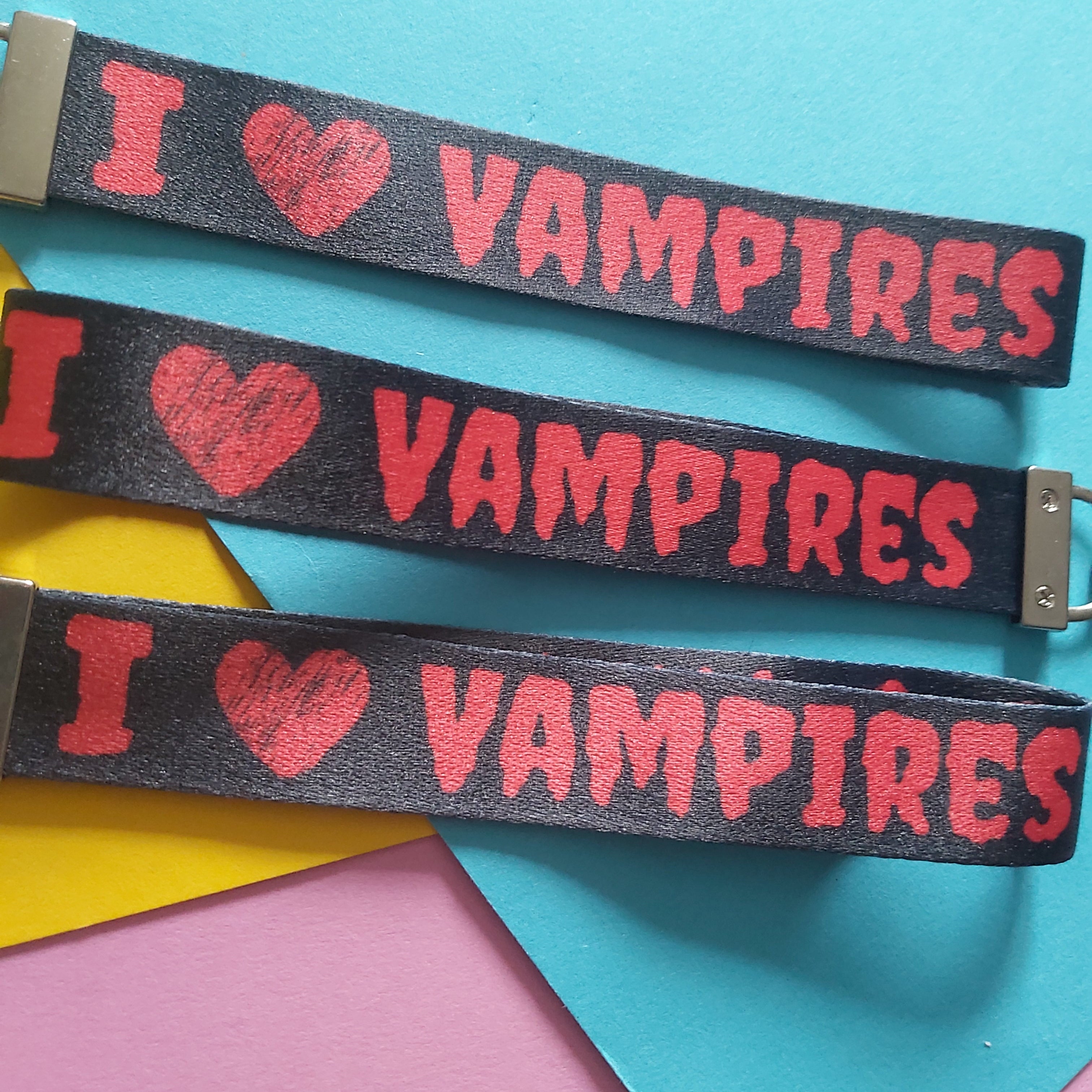 Three black fabric wristbands with silver metal clasps, printed with the red text 'I [heart] VAMPIRES', arranged diagonally on a colorful background of blue, pink, and yellow paper.