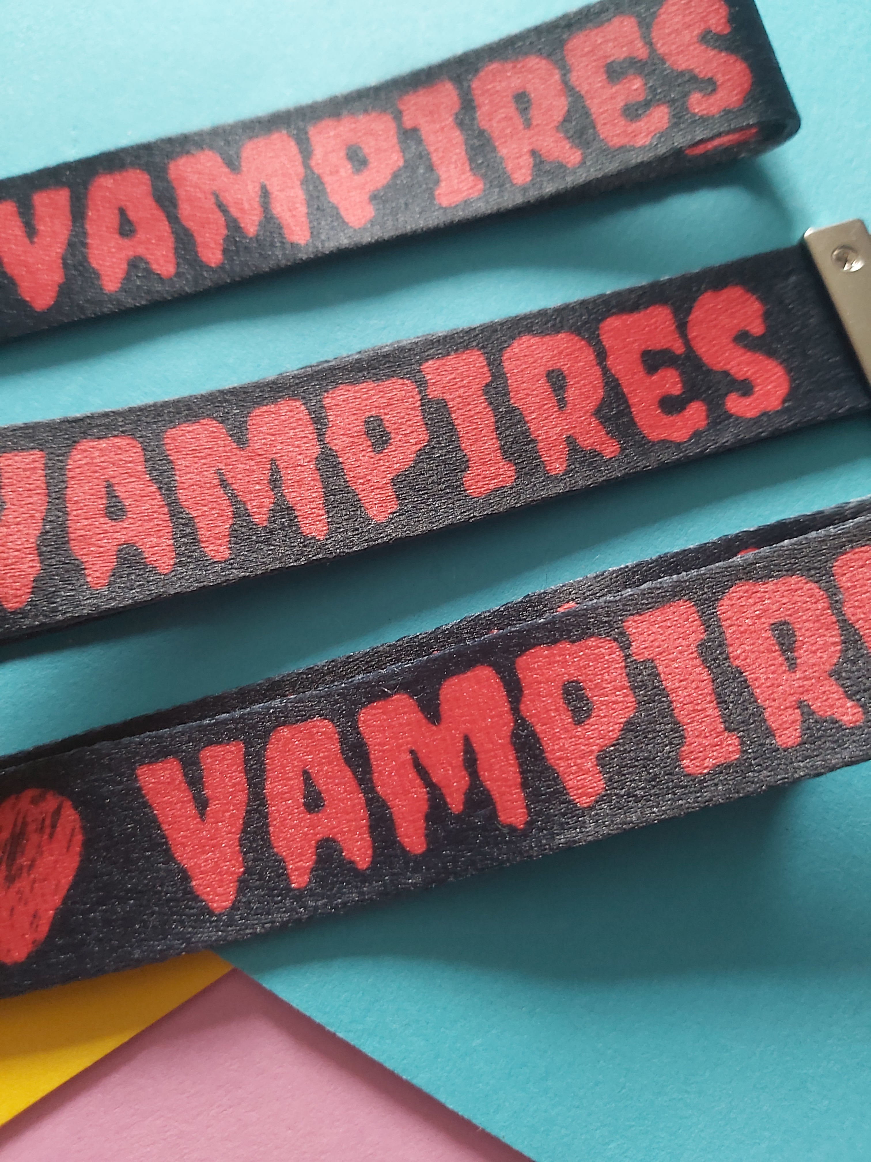 Three black fabric wristbands with red 'VAMPIRES' text, arranged diagonally on a bright teal background with colorful paper accents.