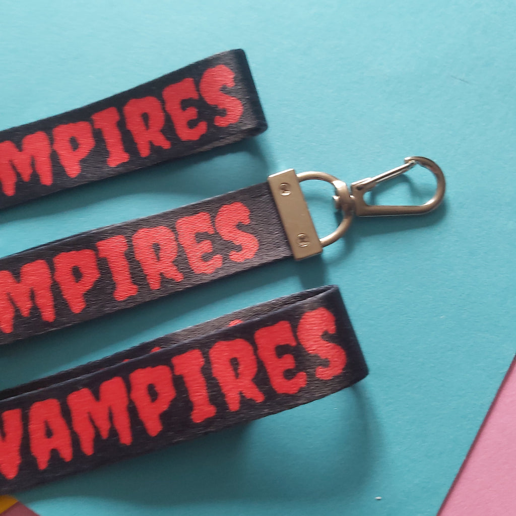 Three black fabric wristlets with red 'VAMPIRES' lettering and silver metal clasp, arranged diagonally on a split turquoise and pink background.