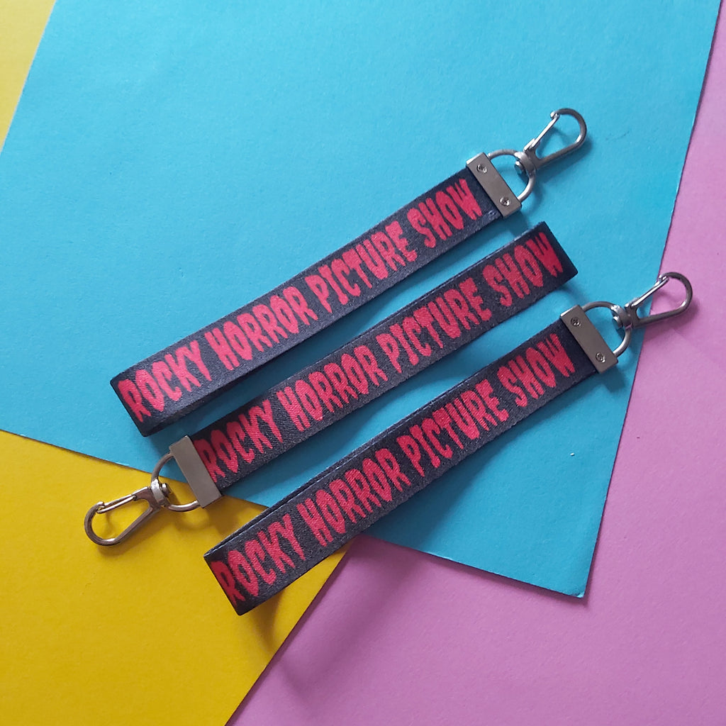 Three black fabric lanyards with silver metal clasp keys, printed with the red text 'ROCKY HORROR PICTURE SHOW', arranged diagonally on a background of colored paper.
