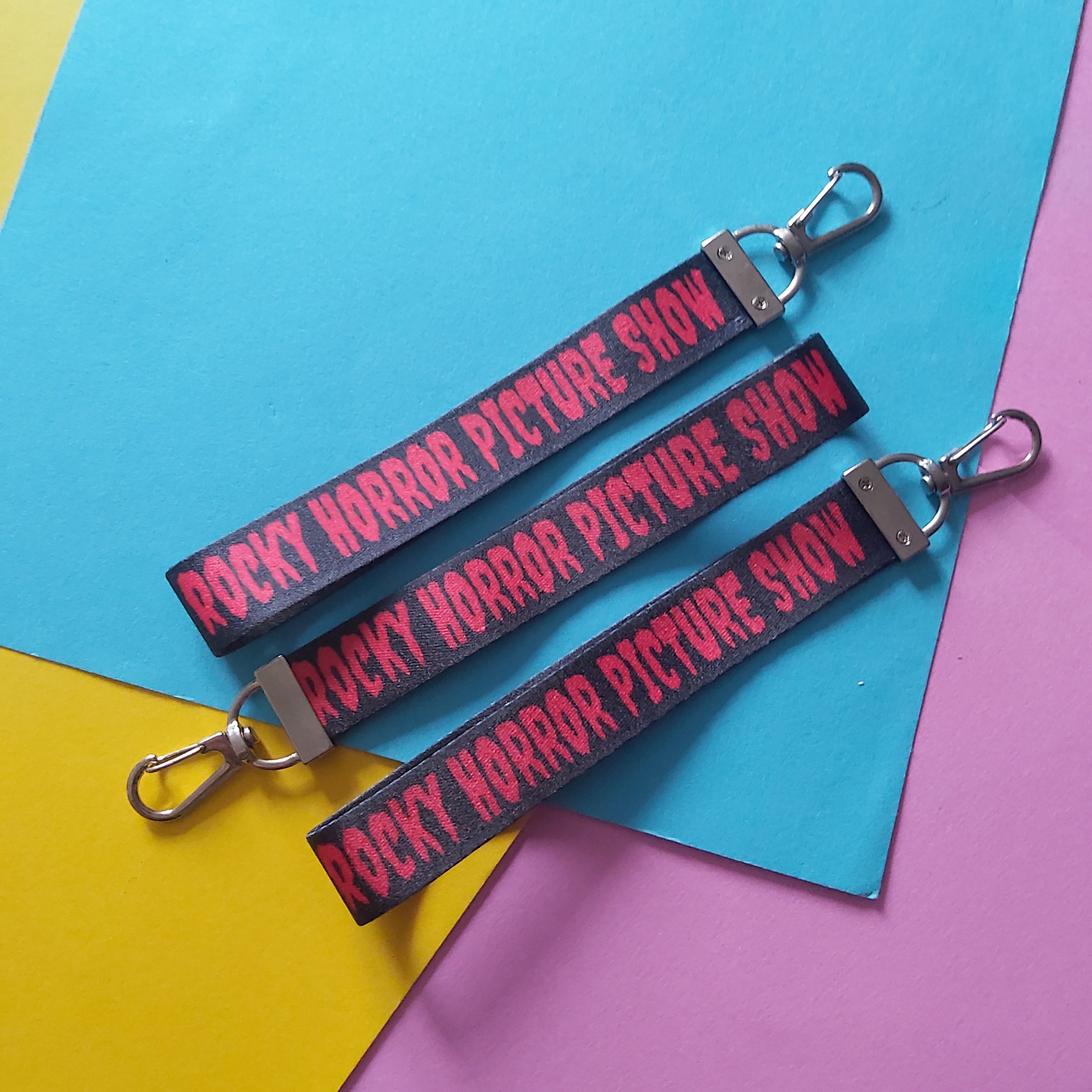 Three black fabric lanyards with silver metal clasp keys, printed with the red text 'ROCKY HORROR PICTURE SHOW', arranged diagonally on a background of colored paper.