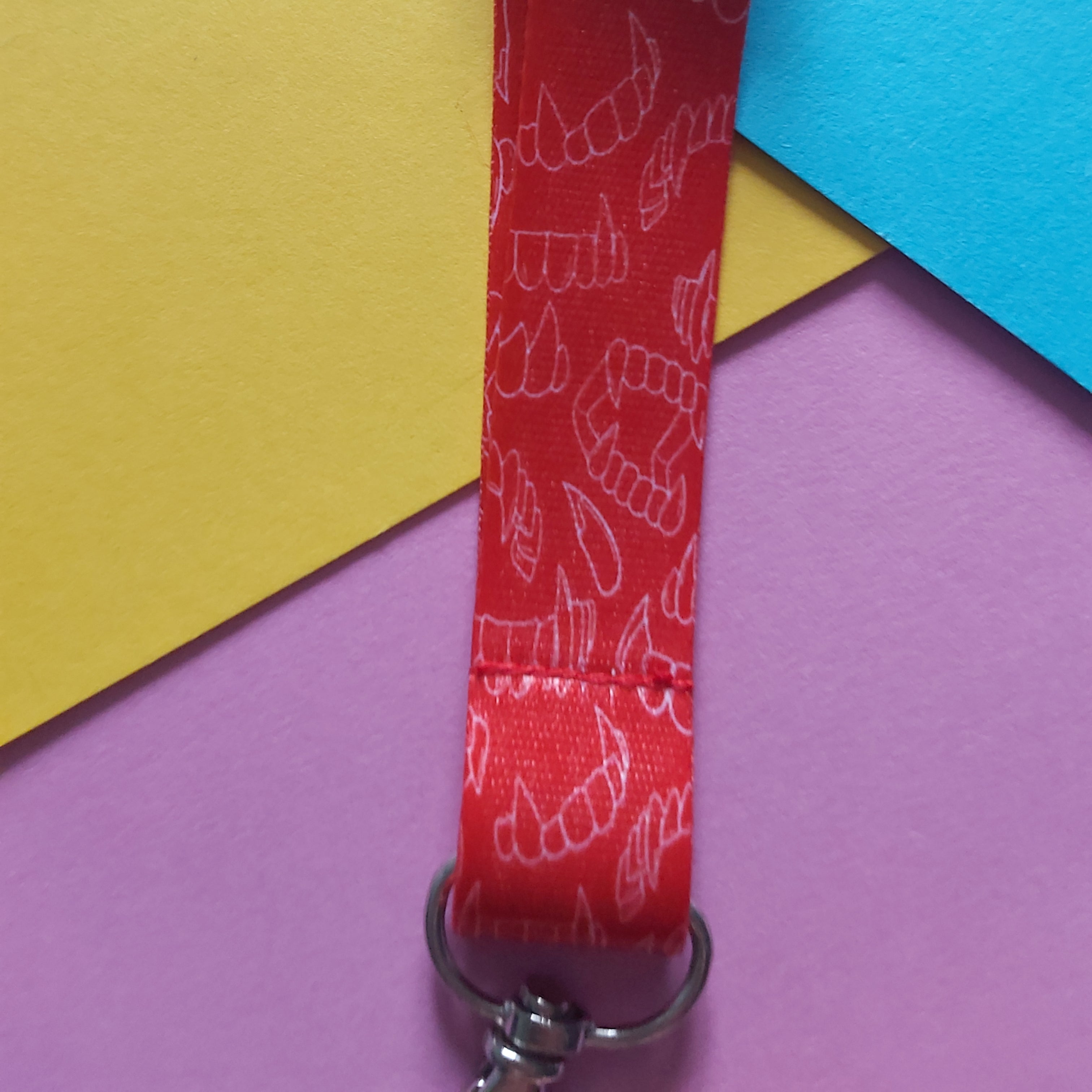 A close-up of a red fabric lanyard with a repeating white line-art pattern of vampire teeth, attached to a silver metal clasp, set against a colourful background of yellow, blue, and purple paper.