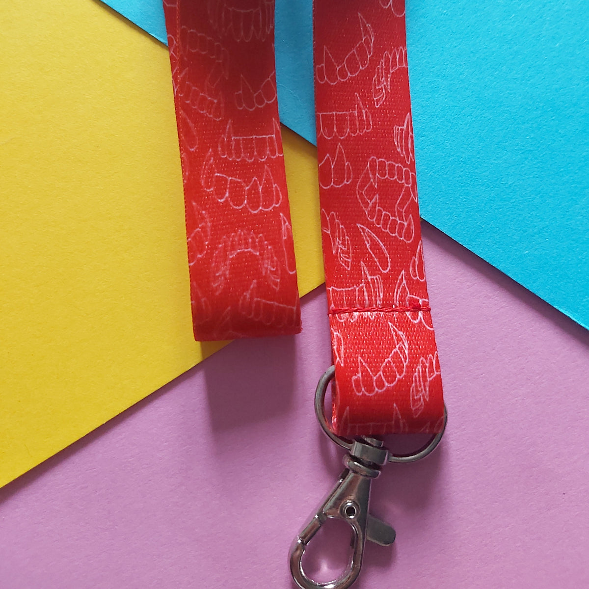 A red fabric lanyard with a repeating white line-art pattern of fangs, attached to a silver metal lobster clasp, laid flat on a colourful background of yellow, blue, and purple paper.