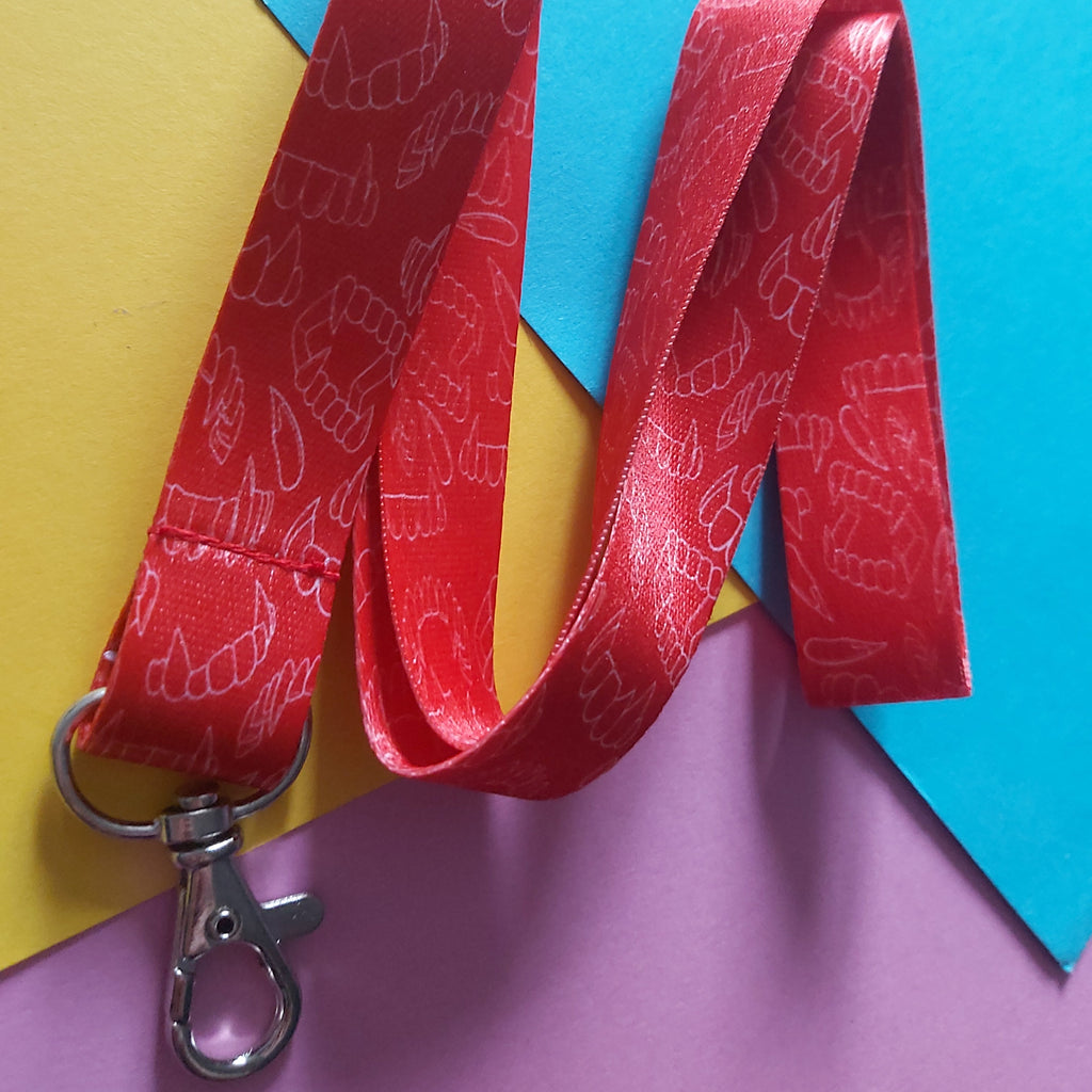 A red cotton lanyard with a repeating white fang pattern, featuring a silver metal lobster clasp, laid out on a colourful background of yellow, blue, and pink paper.