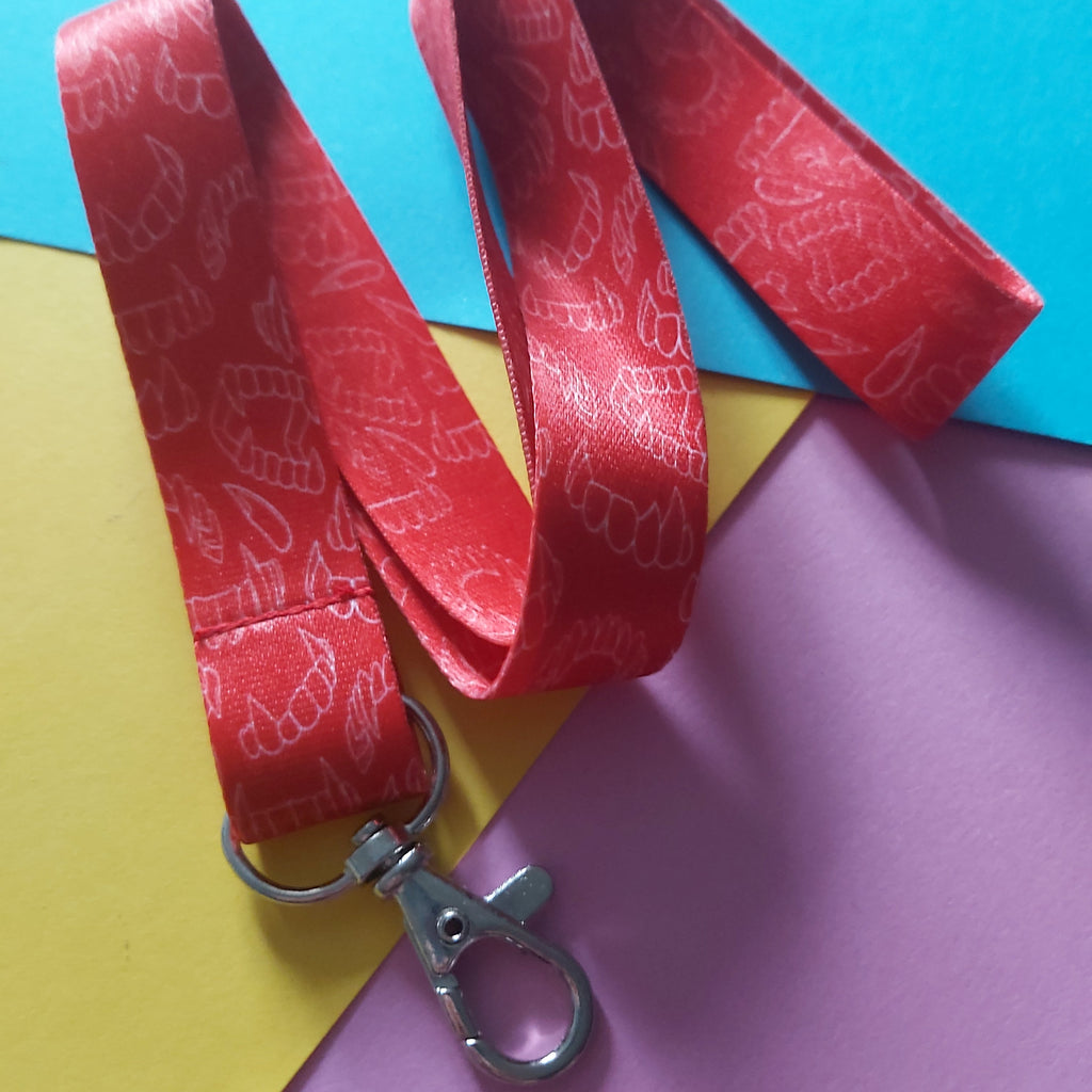 A red fabric lanyard with a repeating white line-art pattern and a silver metal lobster clasp, laid on a colourful background of blue, yellow, and pink paper.