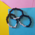 Three beaded bracelets arranged on a bright blue card, with a yellow and pink background. The bracelets are made of glossy black round beads, with three featuring white letter beads spelling 'ATEEZ', 'ENHYPEN' and 'STRAY KIDS' with small black star accents.