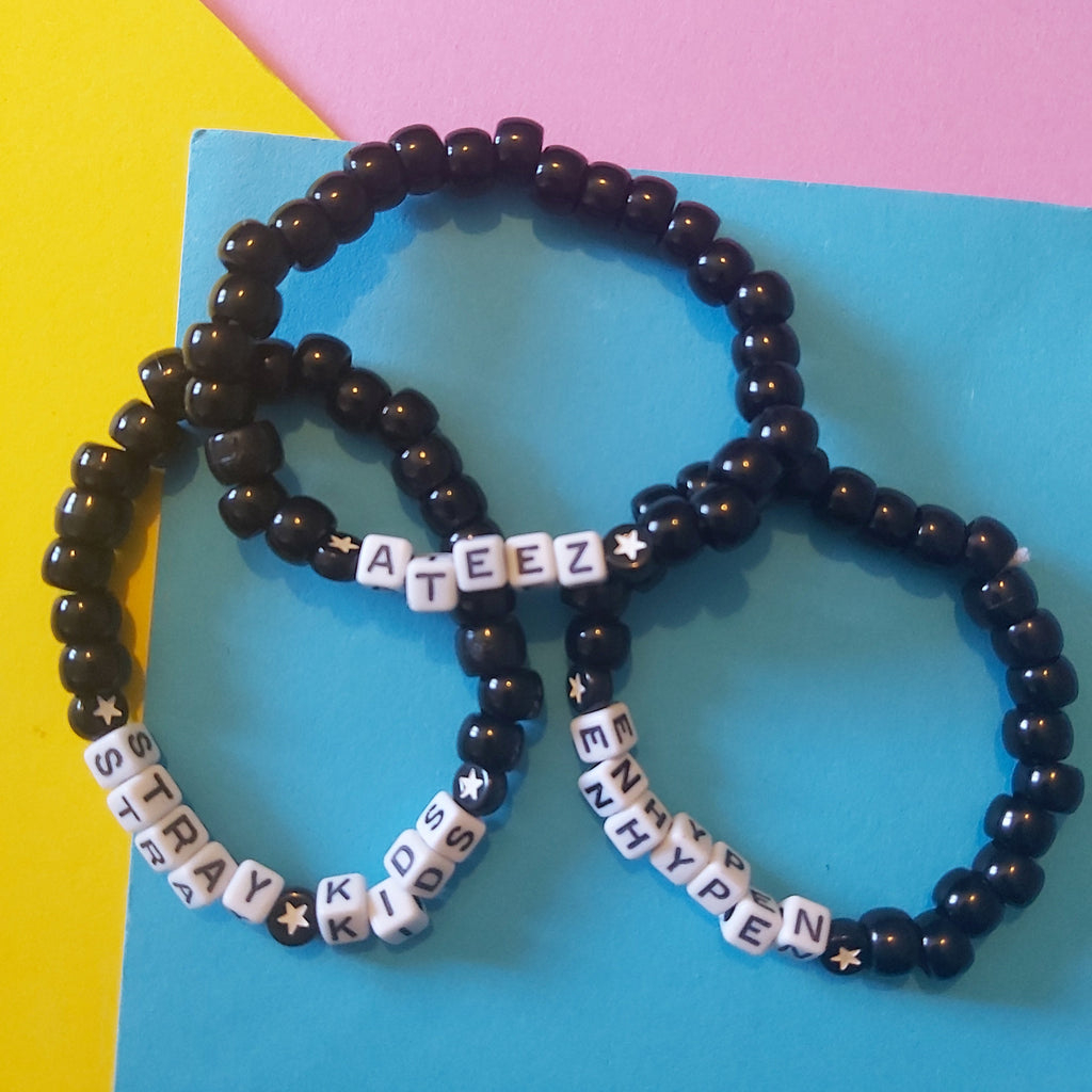 Three beaded stretch bracelets arranged on a bright blue card, with a yellow and pink background. The bracelets are primarily glossy black beads with white letter beads spelling 'ATEEZ', 'STRAY KIDS', and 'ENHYPEN', accented with small white star beads.