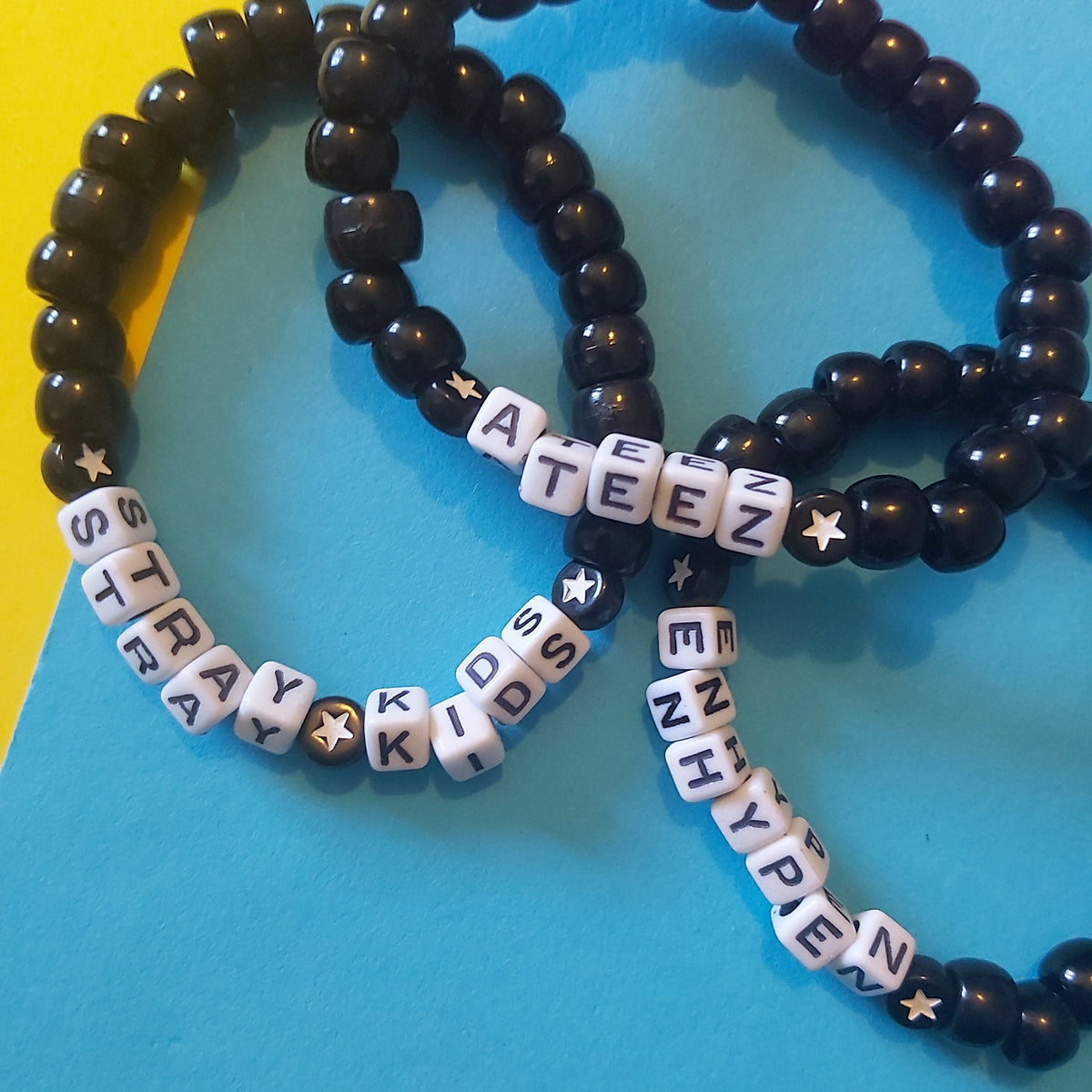 Three beaded stretch bracelets displayed on a colourful background. Two bracelets are made of glossy black beads, and one is made of white letter tiles spelling out kpop groups like 'STRAY KIDS' with small star accents.