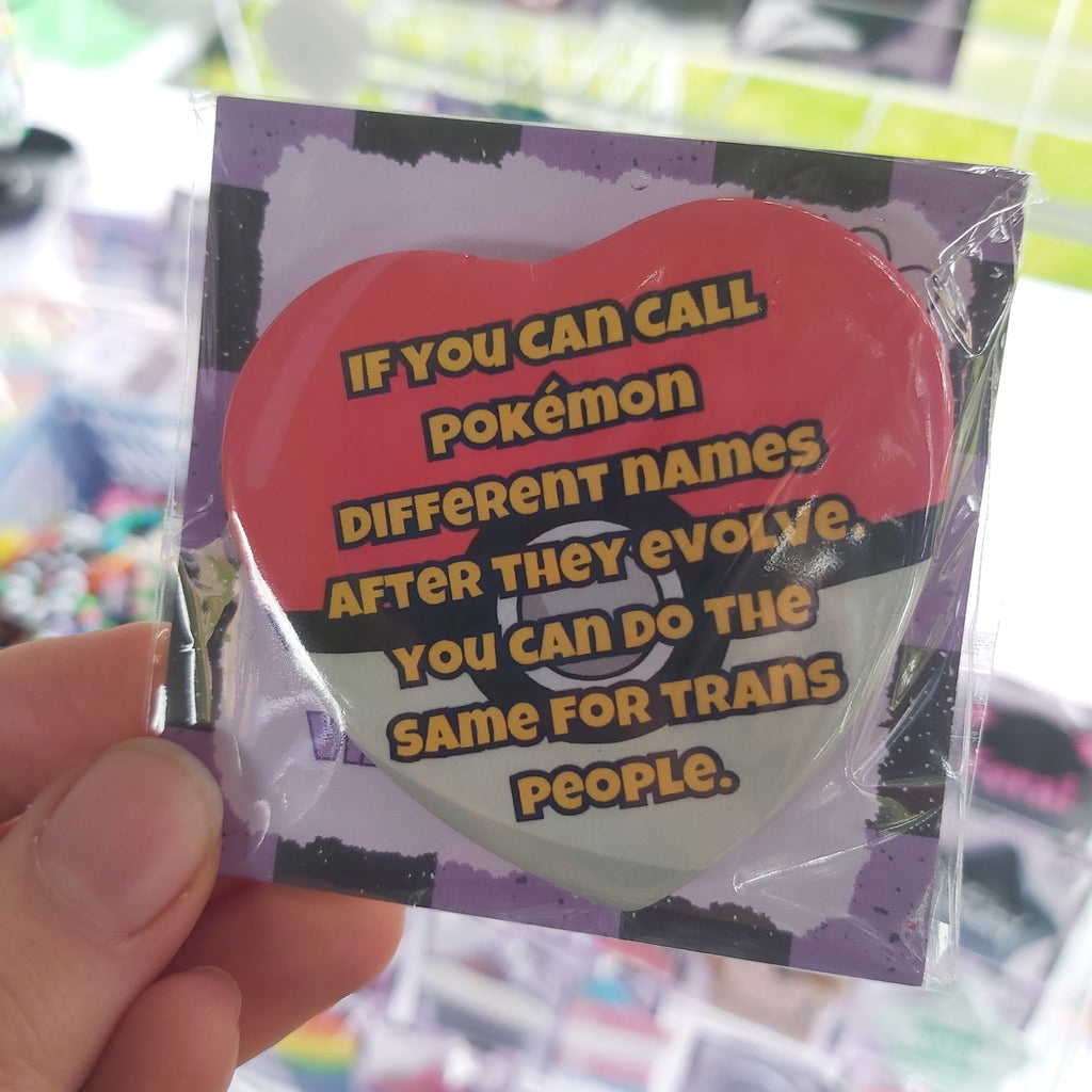 A hand holding a small packaged heart-shaped pin with a red, white, and black design and bold text that reads, 'IF YOU CAN CALL POKEMON DIFFERENT NAMES AFTER THEY EVOLVE. YOU CAN DO THE SAME FOR TRANS PEOPLE.