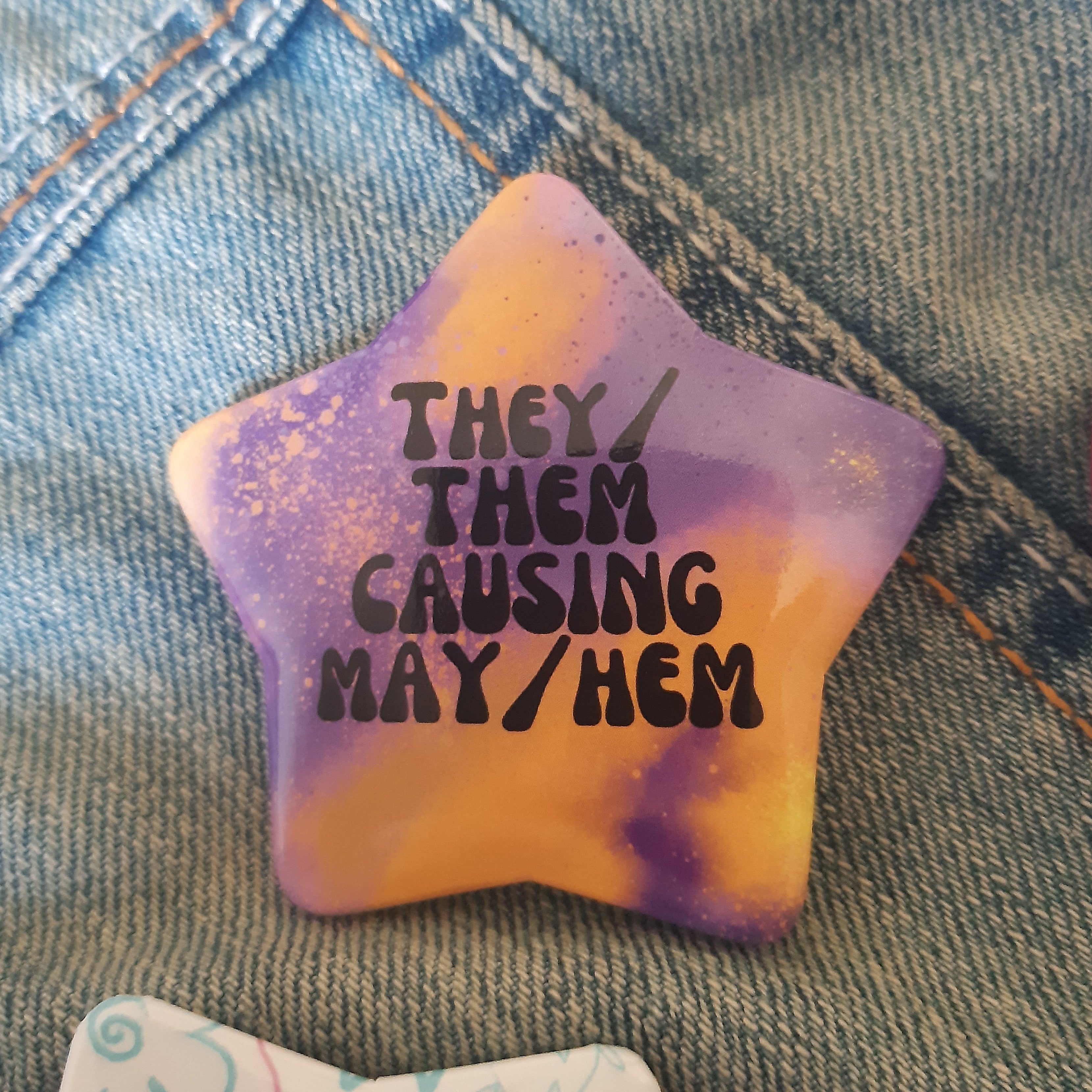 A star-shaped pin with a purple and yellow tie-dye pattern and black text reading 'THEY/ THEM CAUSING MAY/HEM', displayed on a blue denim jacket.