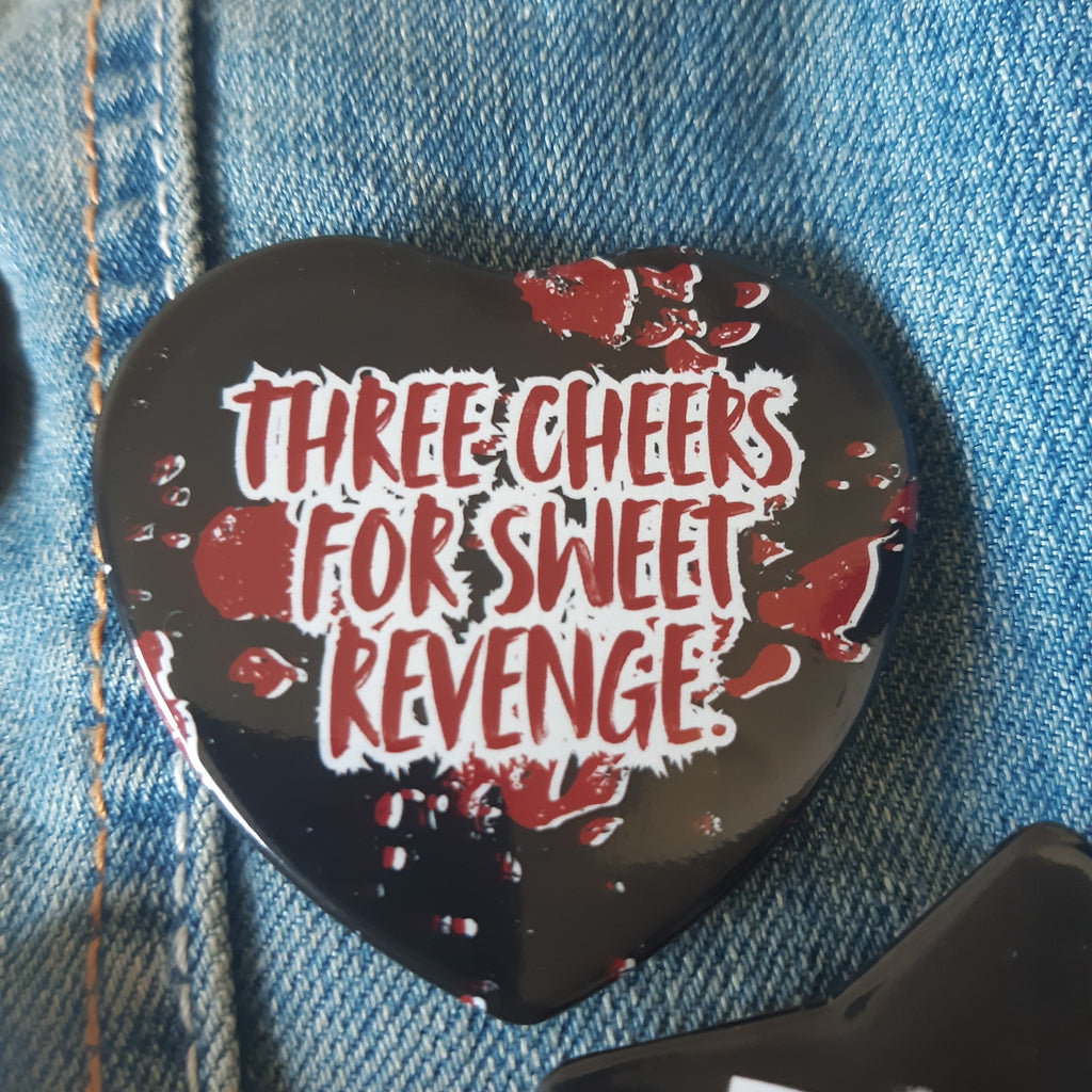 A heart-shaped black badge pin with the phrase 'THREE CHEERS FOR SWEET REVENGE' in white letters and red blood-splatter details, pinned onto a blue denim jacket.