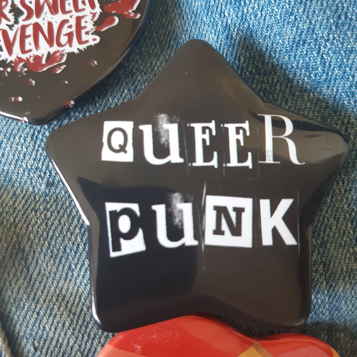 A glossy black star-shaped pin badge with the words 'QUEER PUNK' in white block letters, placed on blue denim fabric next to other colourful pins.
