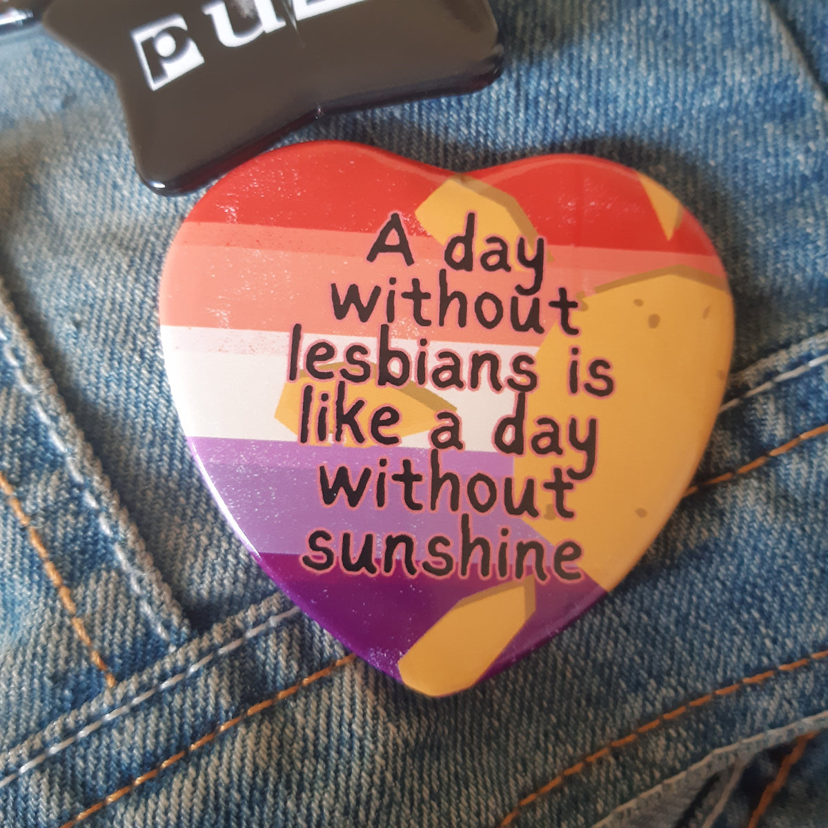 A heart-shaped enamel pin with a rainbow-striped background and the text 'A day without lesbians is like a day without sunshine' in black