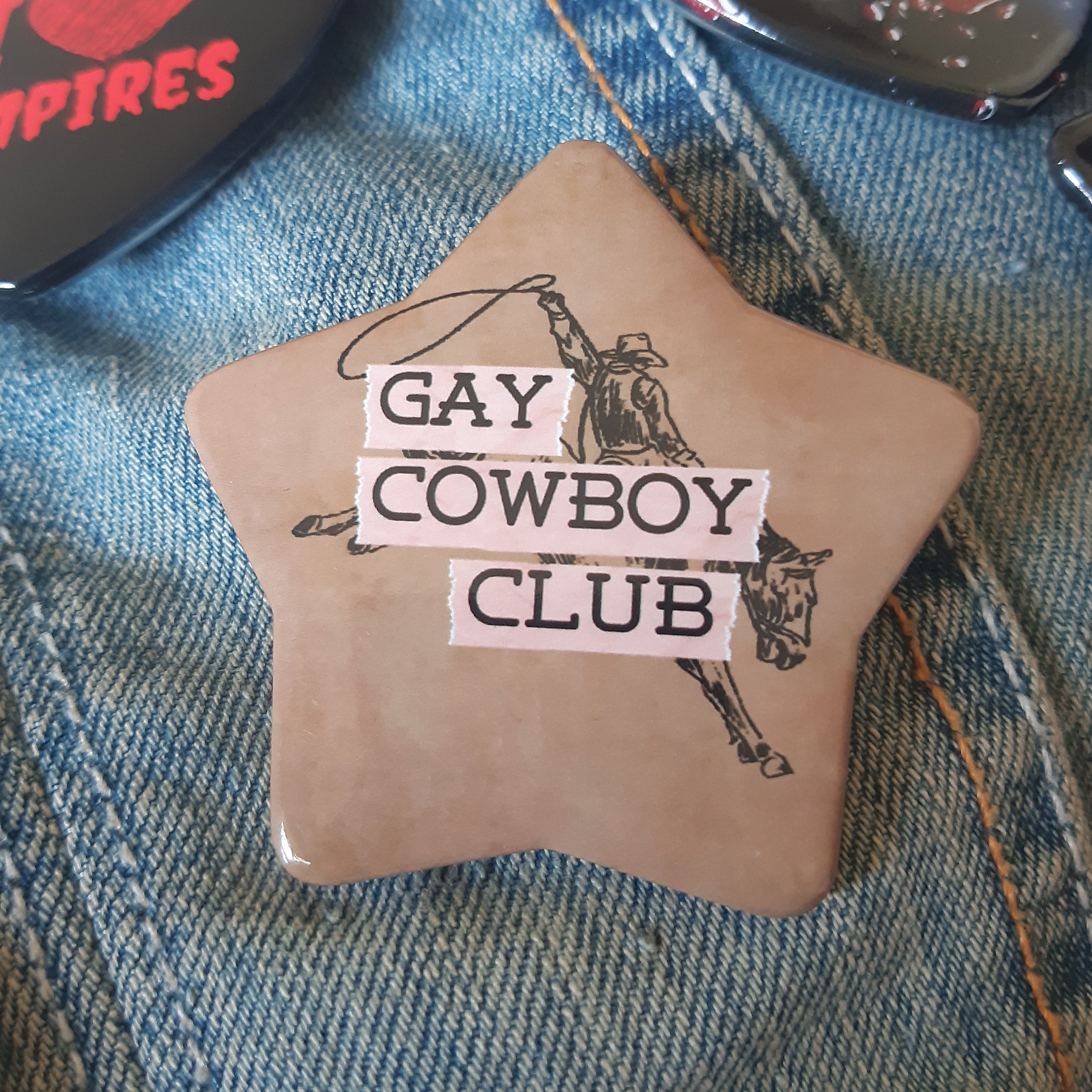 A star-shaped pin badge with a cowboy and lasso graphic, featuring the text 'GAY COWBOY CLUB' in black letters on pink rectangular strips, placed on a blue denim background.