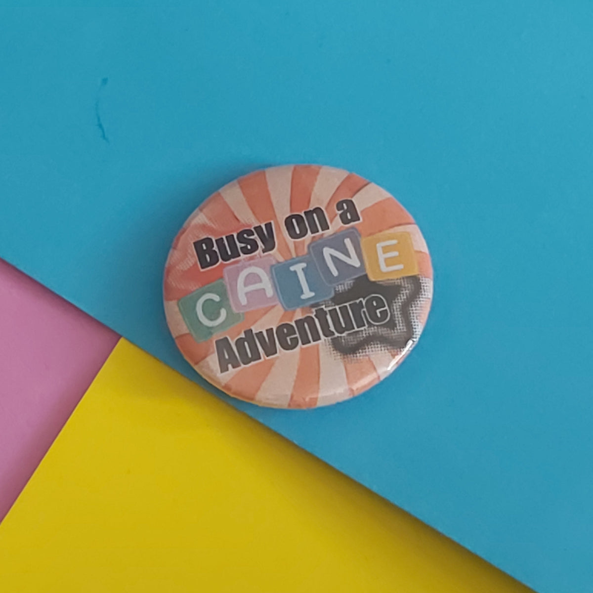 A round pin badge with the text 'Busy on a CAINE Adventure' in black lettering over a retro striped background, placed on a colorful background of blue, yellow, and pink paper.