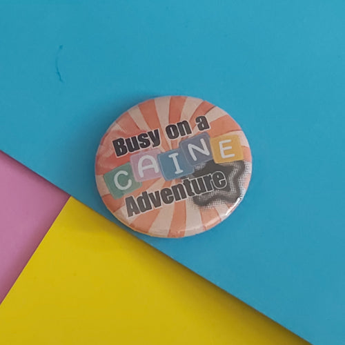 A round pin badge with the text 'Busy on a CAINE Adventure' in black lettering over a retro striped background, placed on a colorful background of blue, yellow, and pink paper.