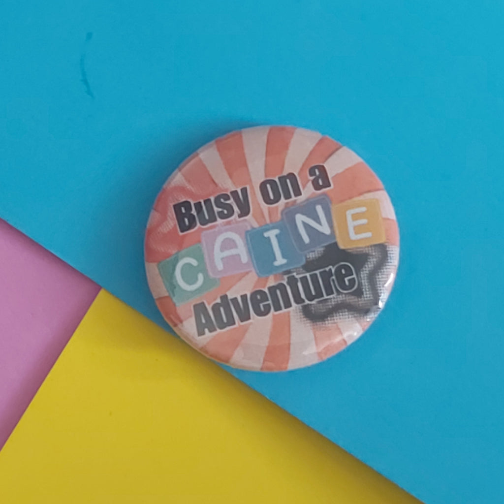 A round pin-back button badge with the text 'Busy on a CAINE Adventure' in black over a pink and orange striped background, placed on a colorful surface with cyan, yellow, and pink sections.