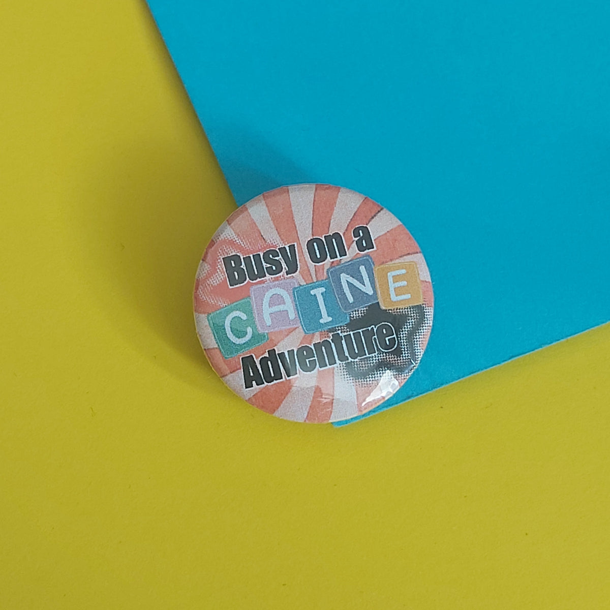 A round pin-back button badge with the text 'Busy on a CAINE Adventure' in black lettering over a pastel striped background, placed on a bright yellow surface with a turquoise paper corner behind it.