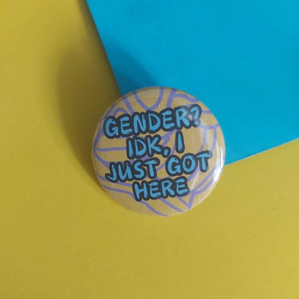 A round pinback button with the text 'GENDER? IDK, I JUST GOT HERE' in blue lettering over a light background with purple scribble lines, placed on a yellow surface with a blue paper corner behind it.