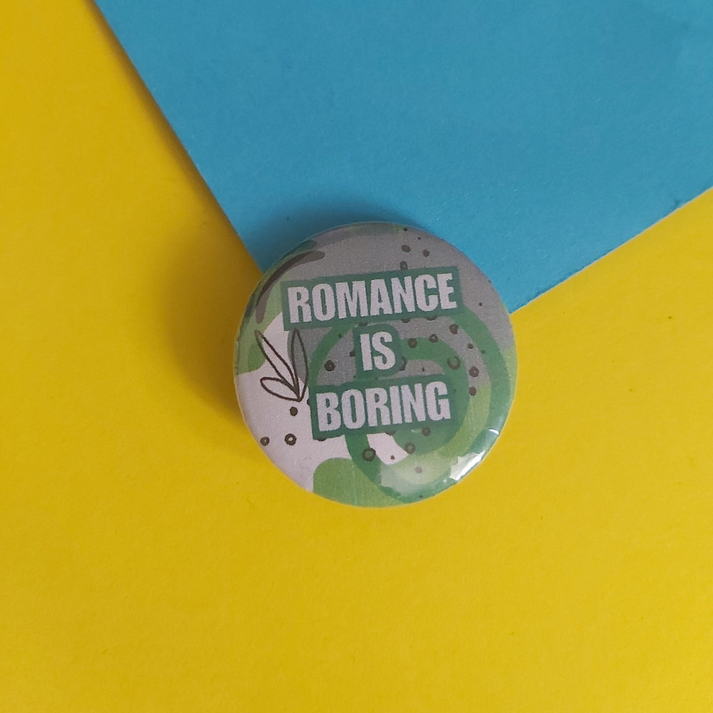 A round pin-back button badge with the text 'ROMANCE IS BORING' in white letters over a green and grey abstract pattern, placed on a split yellow and blue background.