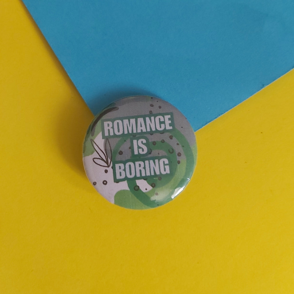 A round pin-back button badge with the text 'ROMANCE IS BORING' in white on a gray background with green leaf illustrations, placed on a split yellow and blue paper backdrop.