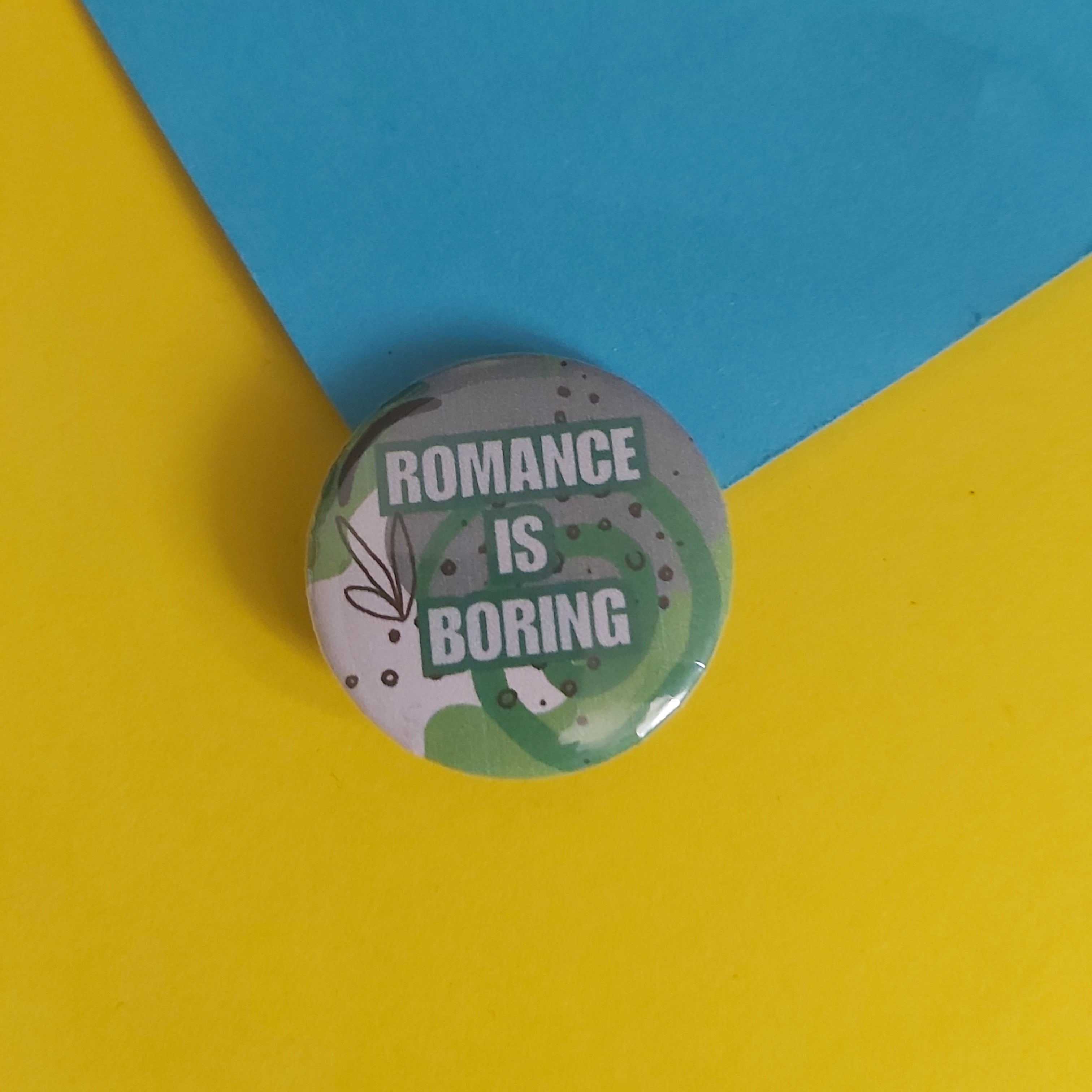 A round pin-back button badge with the text 'ROMANCE IS BORING' in white on a gray background with green leaf illustrations, placed on a split yellow and blue paper backdrop.