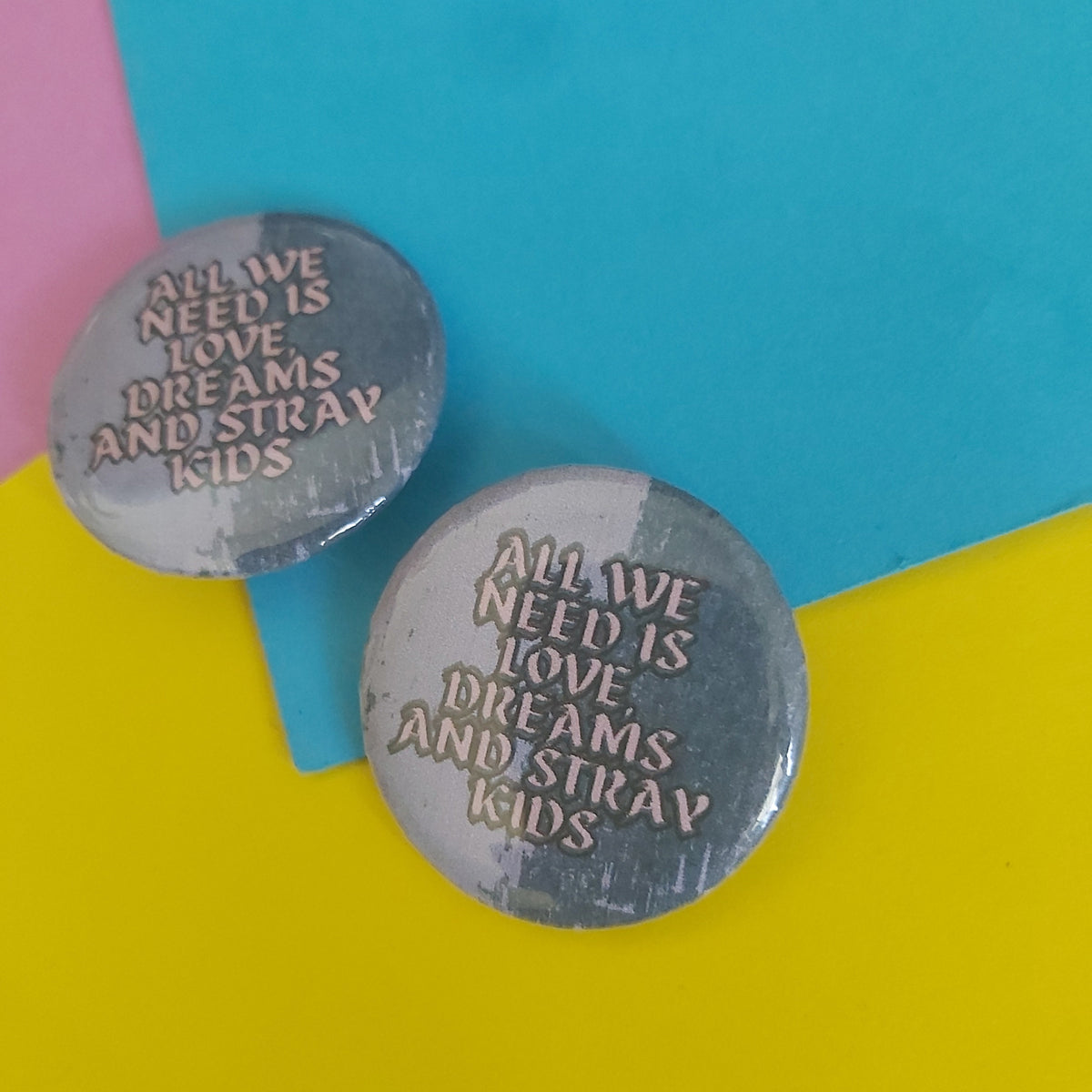 Two round pin-back buttons with the text 'ALL WE NEED IS LOVE DREAMS AND STRAY KIDS' in pink, displayed on a colourful background of yellow, blue, and pink paper.