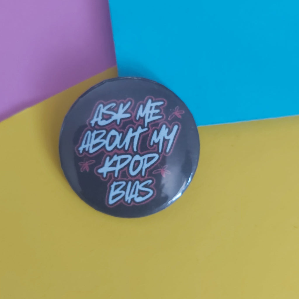 A round pin badge with the text 'ASK ME ABOUT MY K-POP BIAS' in stylized redand white lettering on a dark grey background, photographed on a colourful background of yellow, blue, and pink paper.