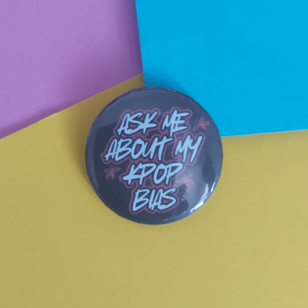 A round pin-back button badge with a dark background and stylised text reading 'ASK ME ABOUT MY K-POP BIAS' in light blue and pink, placed on a yellow surface with purple and blue geometric paper shapes behind it.