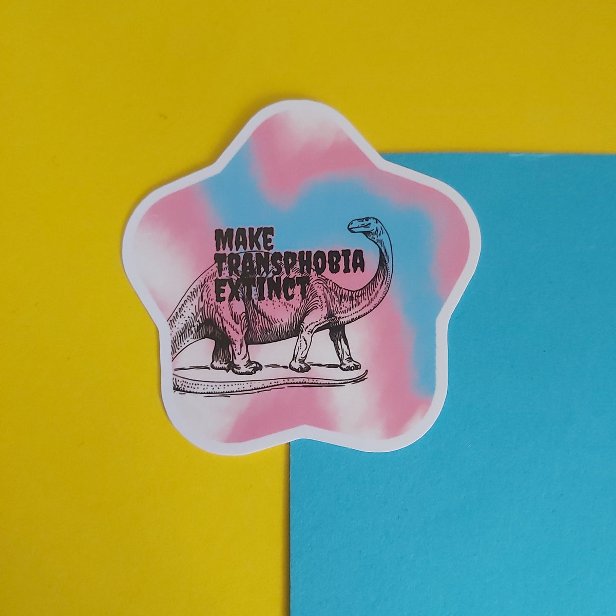 A die-cut sticker featuring a pink and blue tie-dye background with a black line drawing of a long-necked dinosaur and the text 'MAKE TRANSPHOBIA EXTINCT'. The sticker is displayed on a split yellow and blue background.