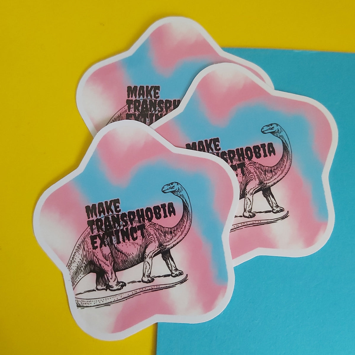 Three die-cut stickers with a pink and blue tie-dye background, featuring an illustration of a dinosaur and the text 'MAKE TRANSPHOBIA EXISTENT', arranged on a split yellow and blue background.